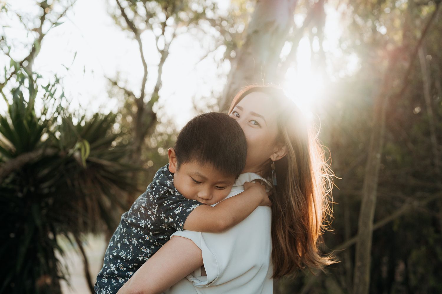 A mother and child share tender moments in a sunlit outdoor setting with trees and natural scenery.