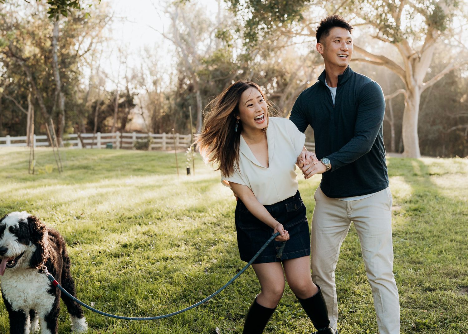 A couple playfully runs and dances together in a sunlit park with their dog during a romantic outdoor photo session.