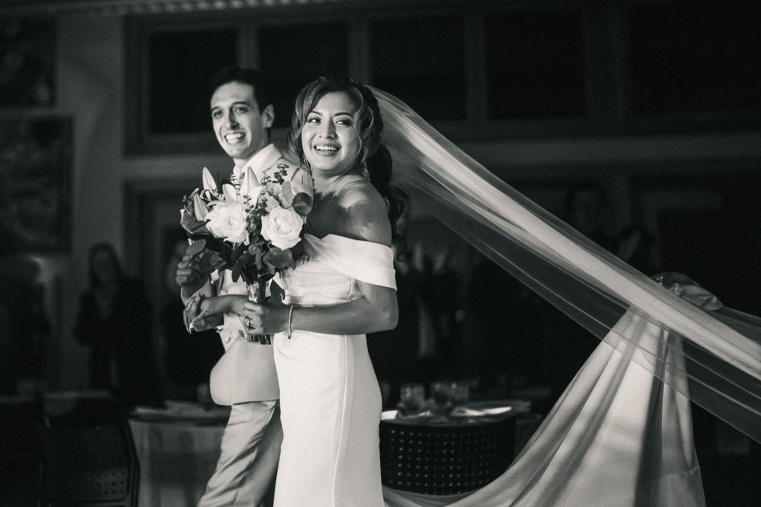 A joyful wedding moment captured in black and white with flowing veil and floral bouquet details.
