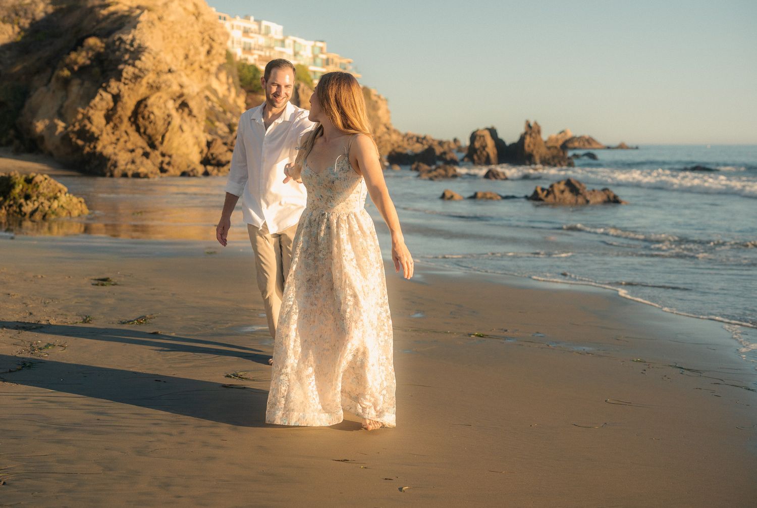 Romantic beach photoshoot at sunset with rocky cliffs and ocean waves creating a dreamy coastal backdrop.