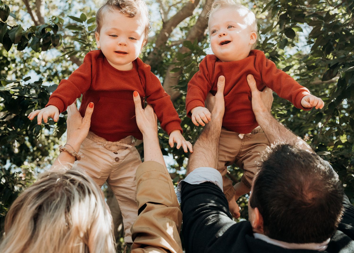 Two toddlers in matching red sweaters and khaki pants being lifted up outdoors among green foliage.