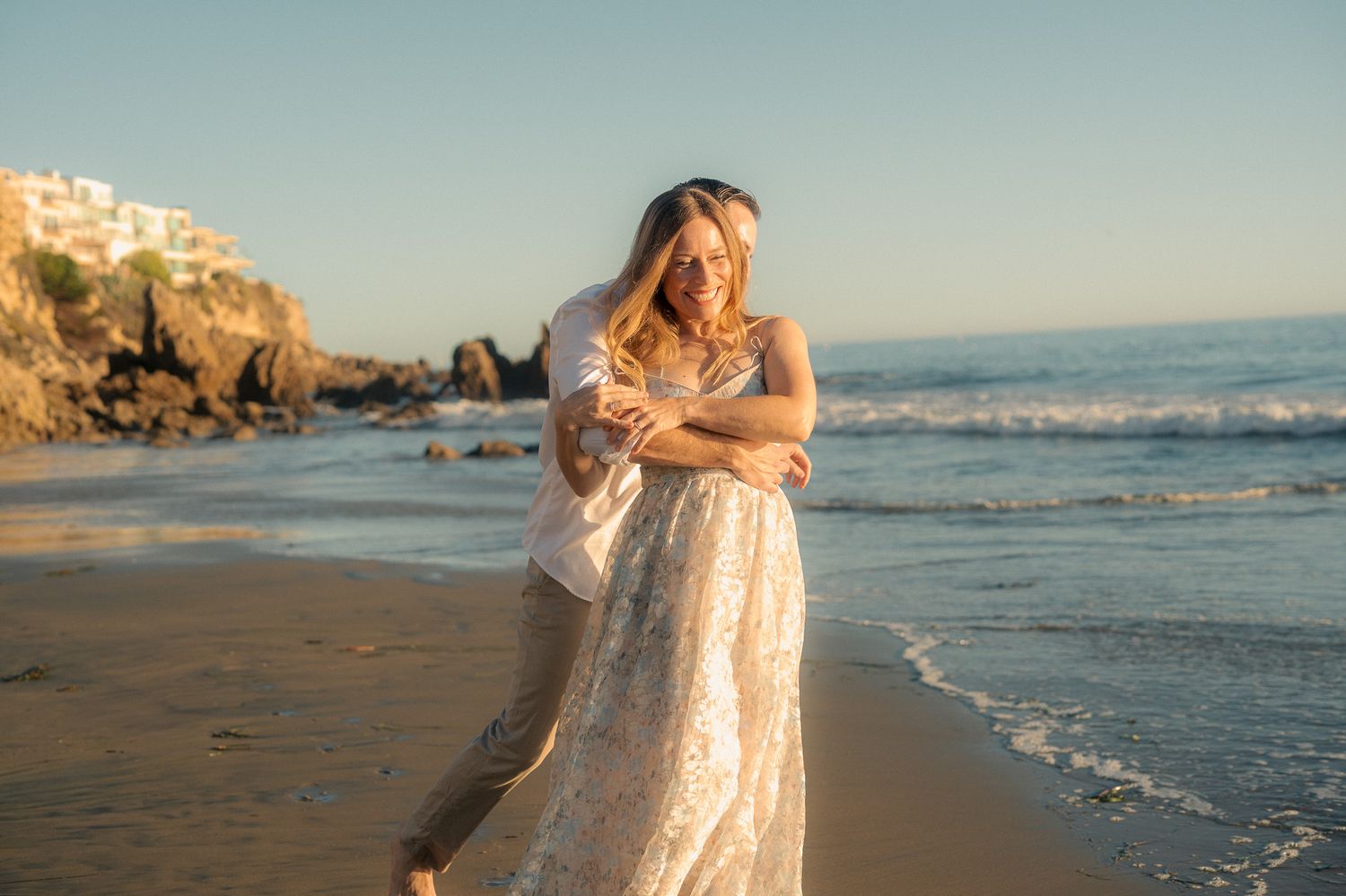 A person in a flowing white dress walks along a scenic beach at sunset with waves crashing in the background.