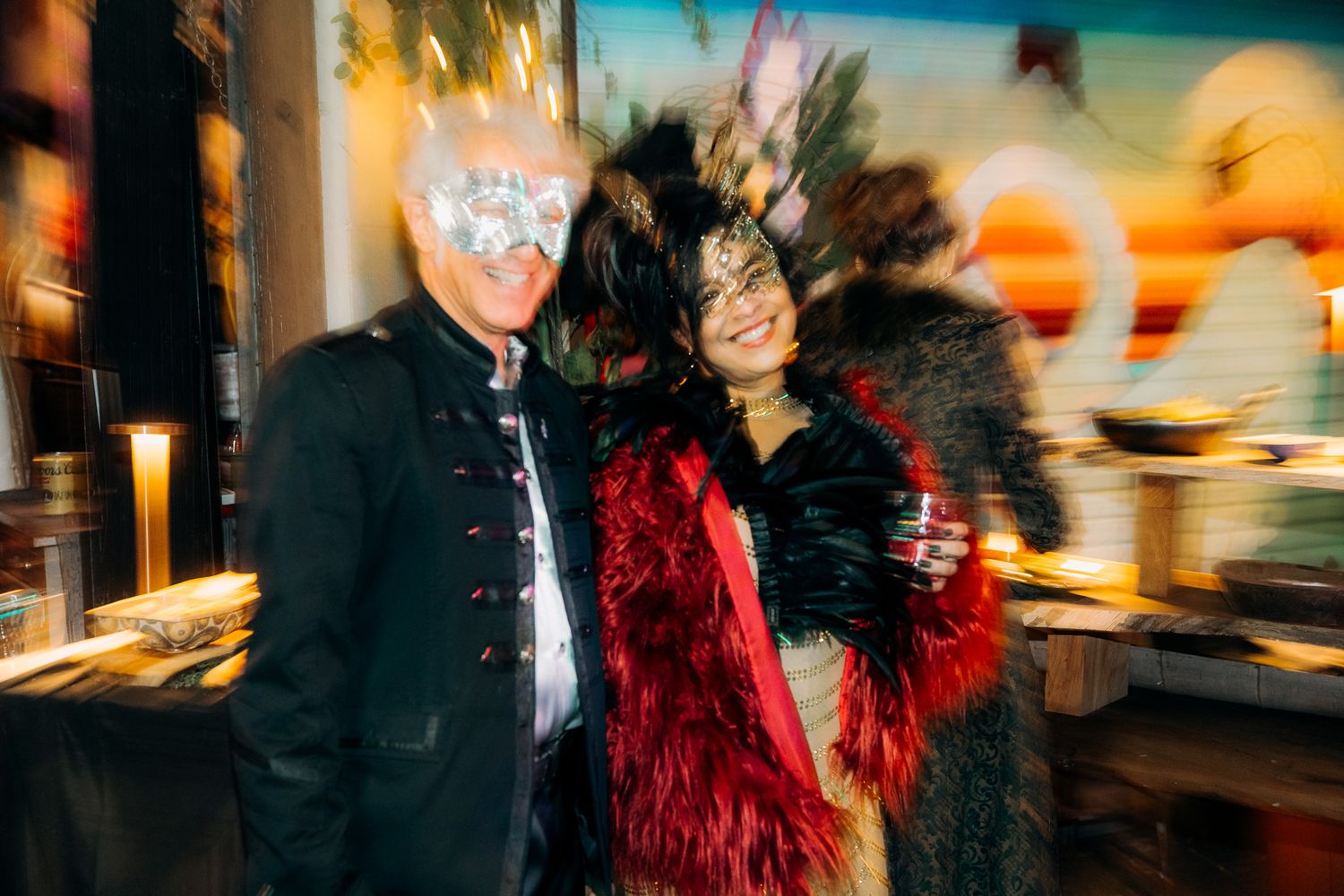 A person wearing a red feathered jacket stands in front of blurred city lights at night.