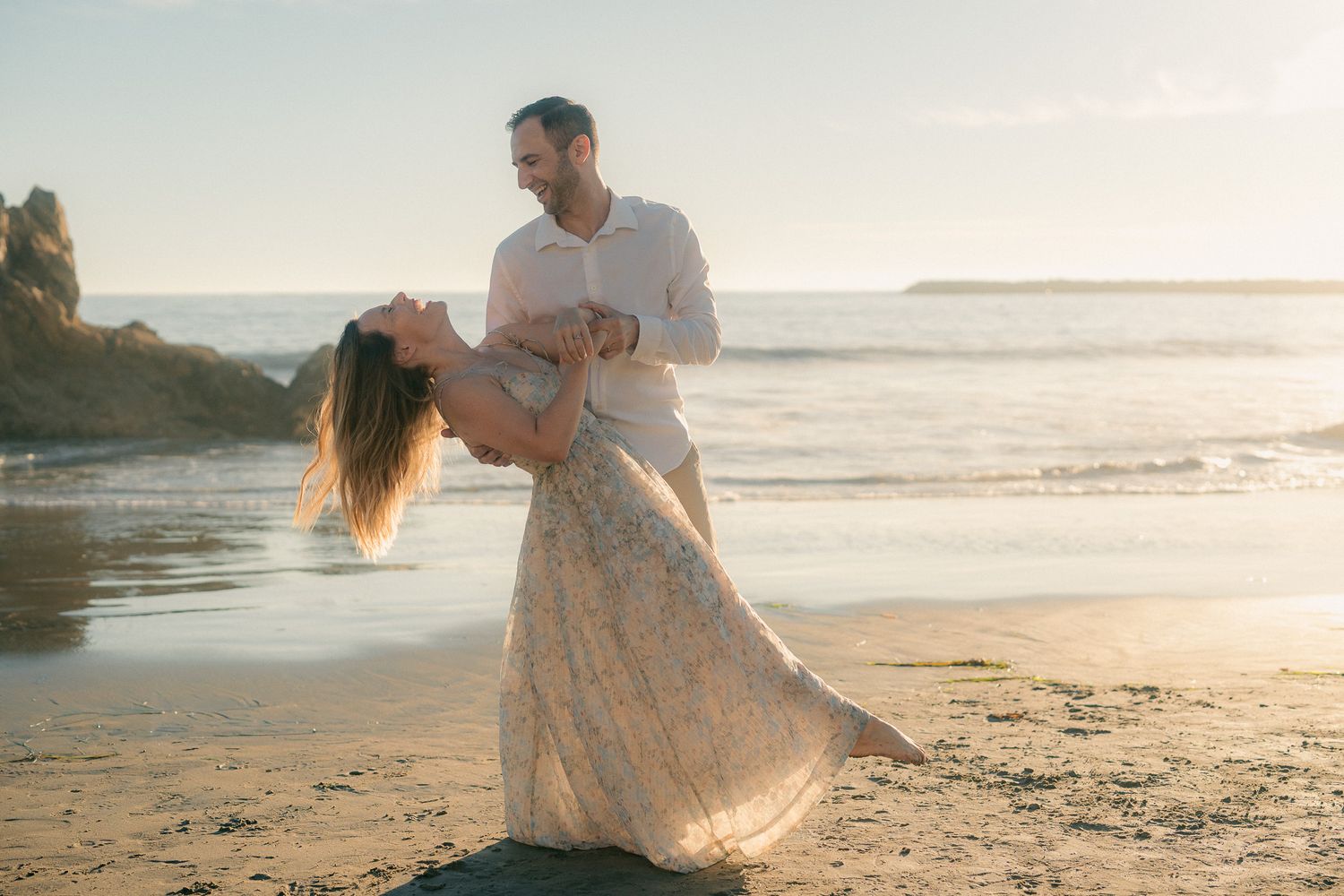 A romantic couple embraces on a sandy beach at sunset creating silhouettes against the golden sky.