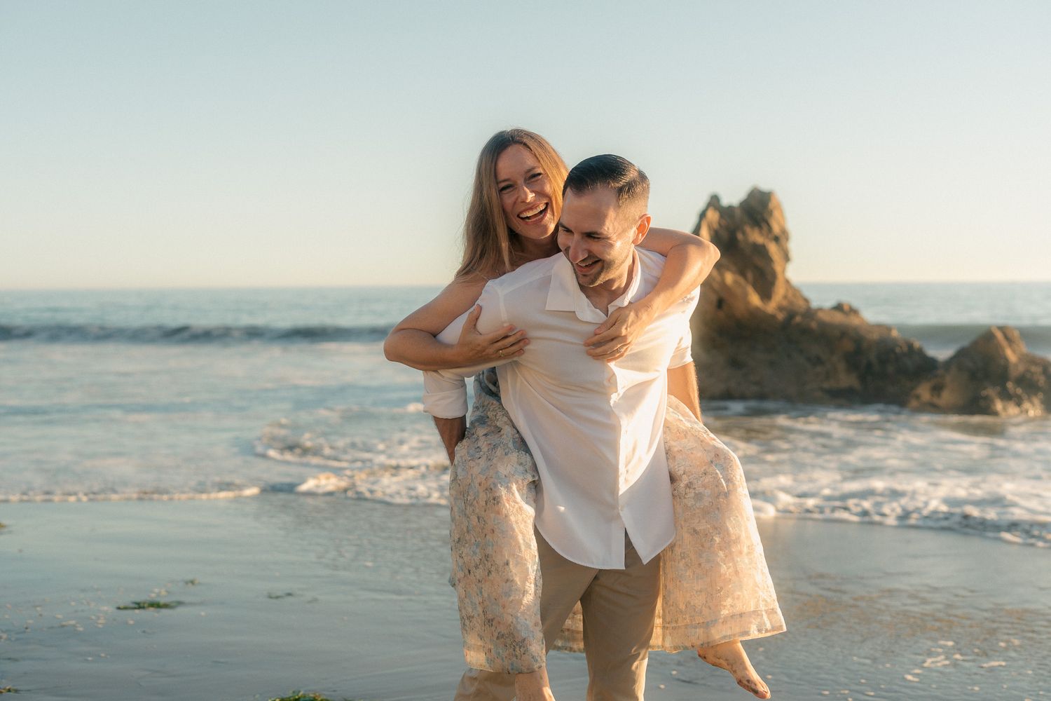 A couple in light-colored outfits shares a romantic moment together on a sunny beach at sunset.