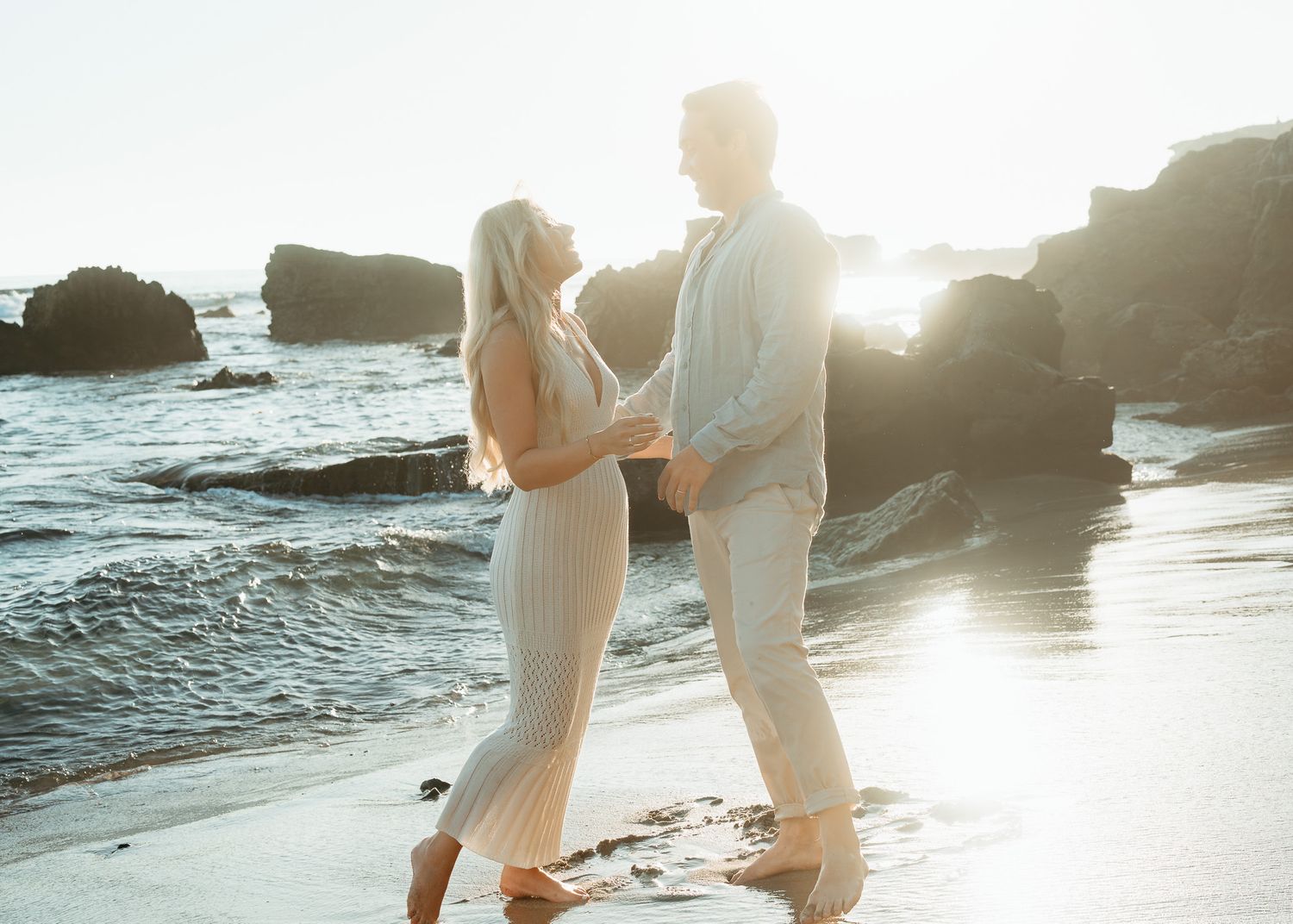 Couple walking along sandy beach at golden hour with dramatic rock formations and crashing waves.
