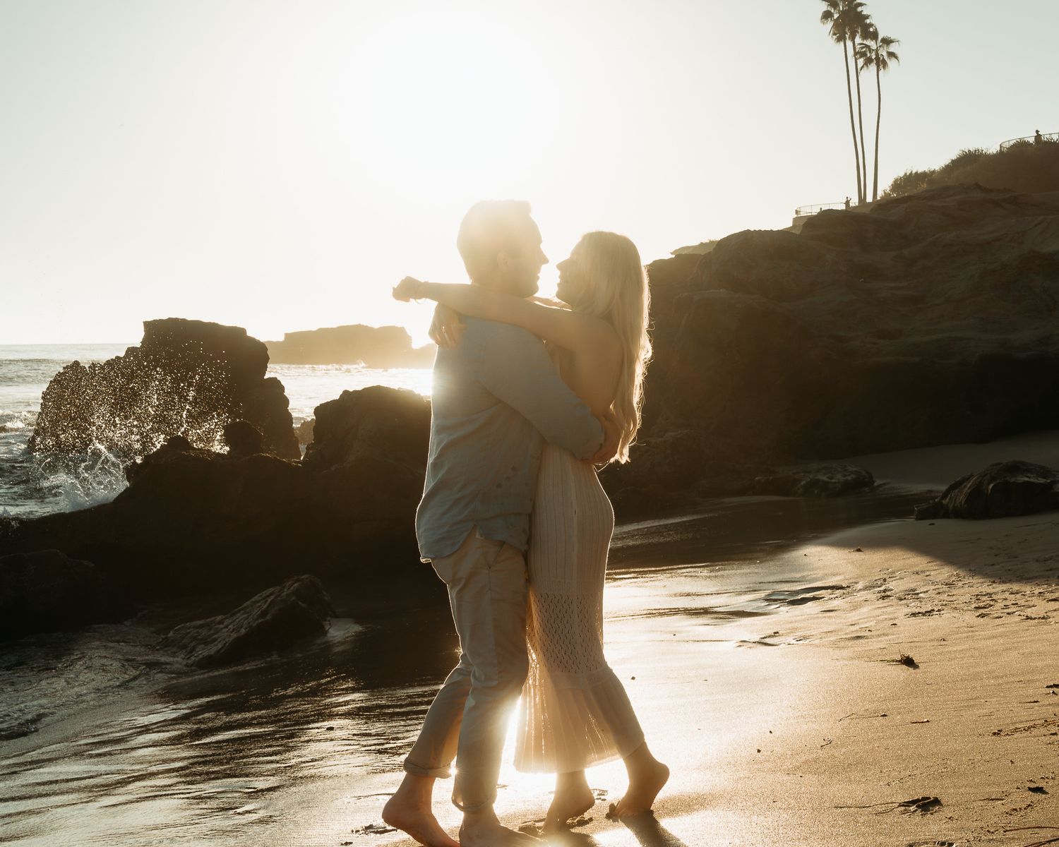 Silhouetted figures walk along a scenic beach at sunset with rocky cliffs in the background.