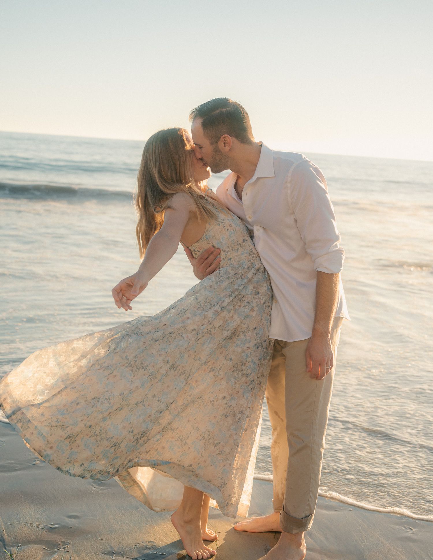 Romantic couple sharing intimate moments on a beach at sunset with flowing floral dress.