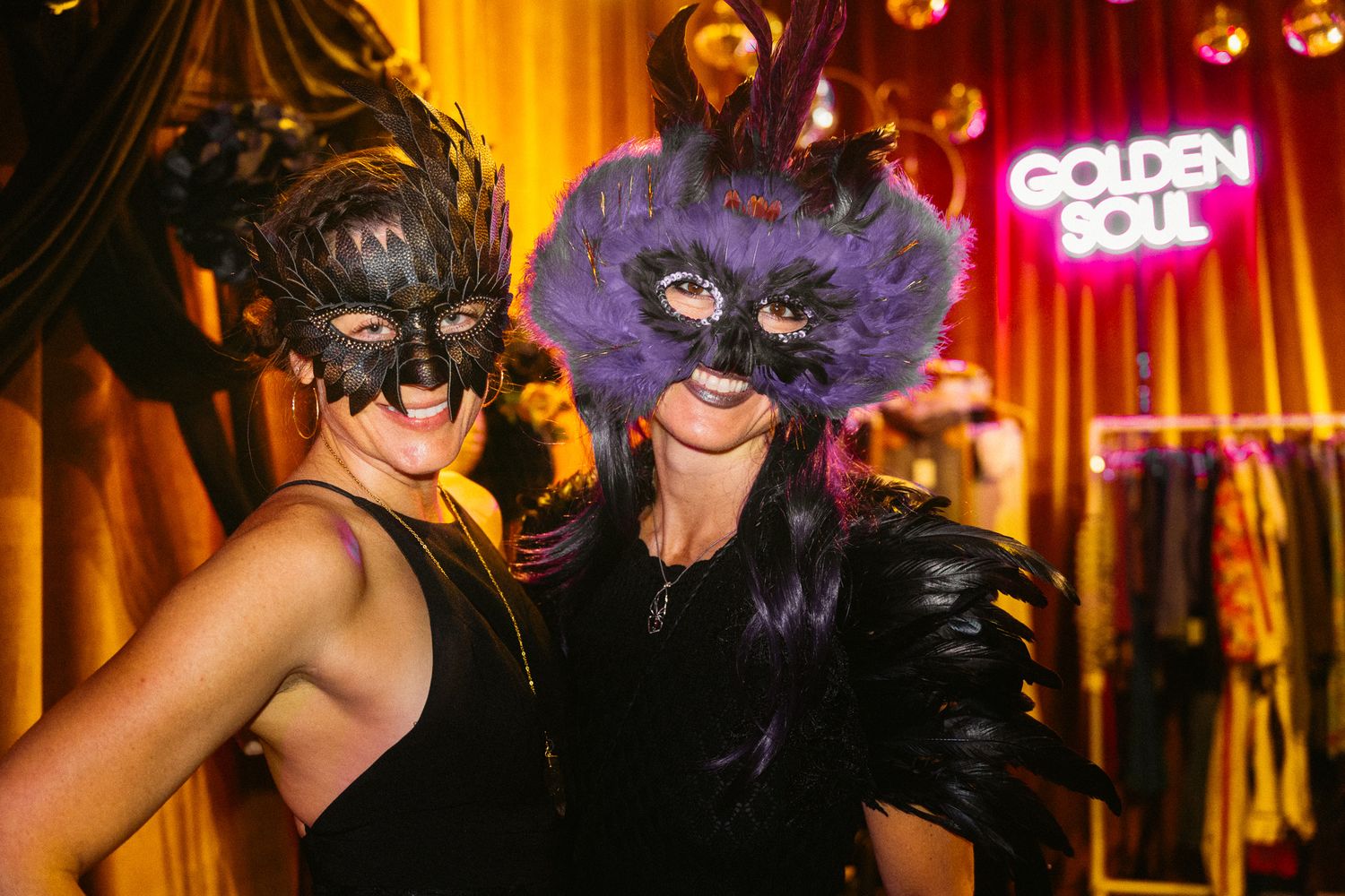 People in black and purple masquerade masks pose together at a glamorous evening event with neon signage.