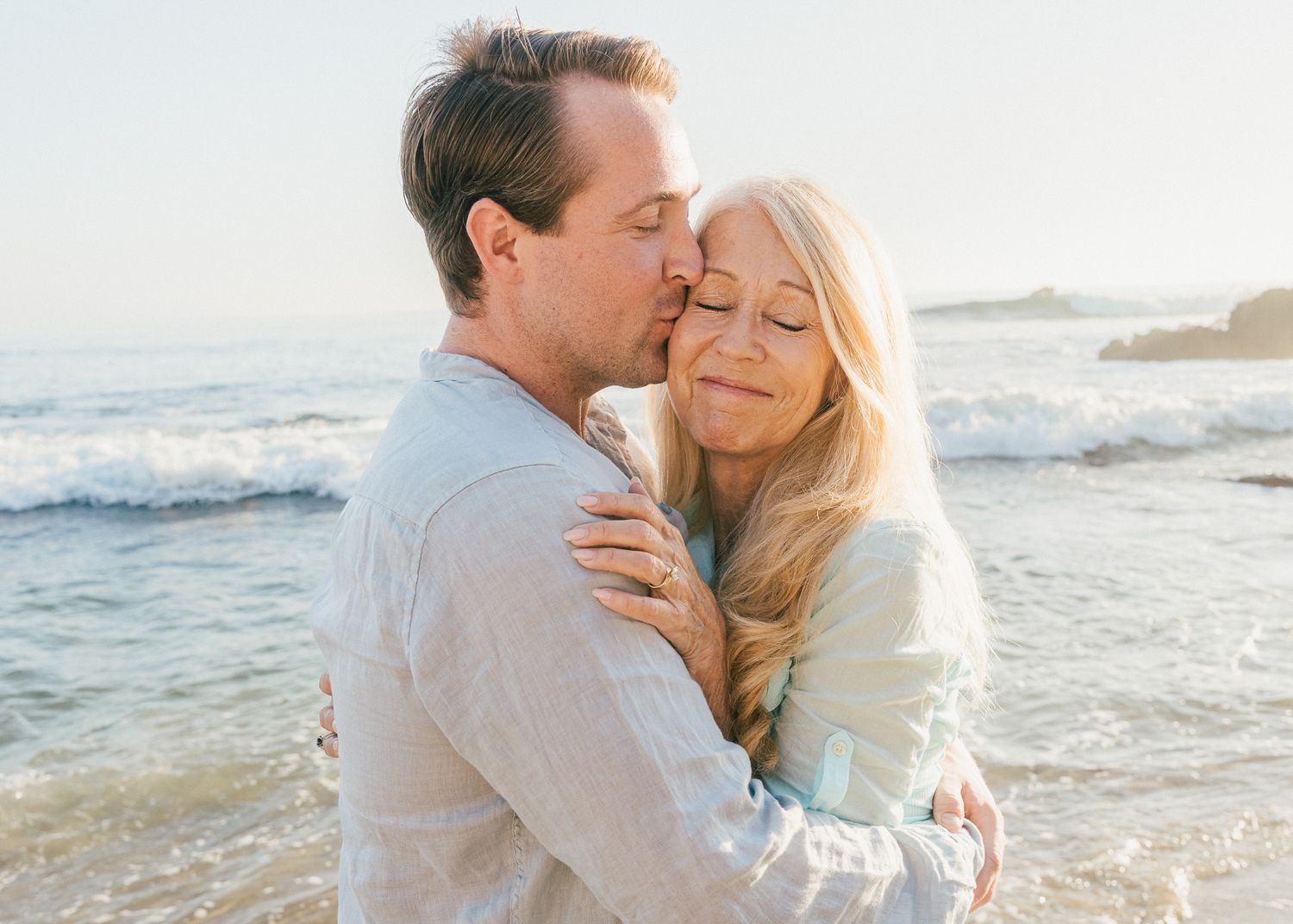 A romantic couple embraces on a sunny beach at sunset with ocean waves in the background.
