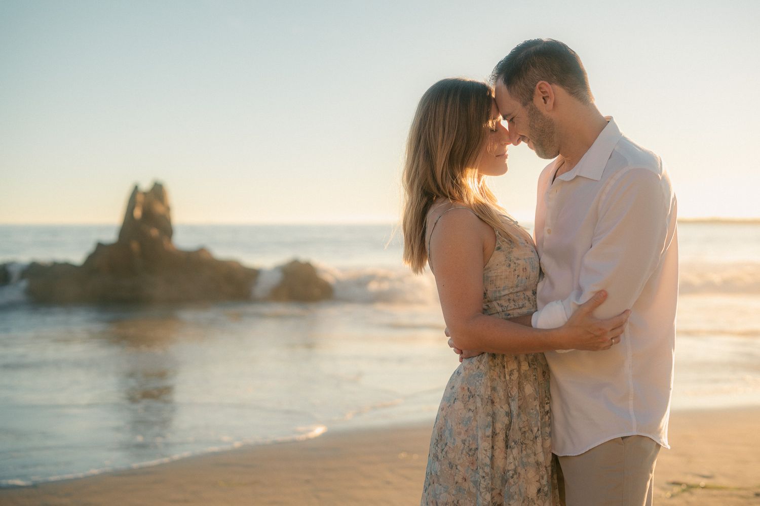 Couple embracing on a scenic beach with rocky coastline and ocean waves at sunset.