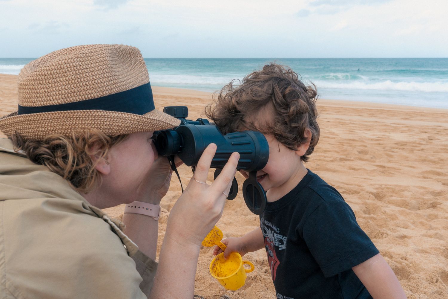 A child and mother playing with binoculars while sitting on sandy shores. Mariano Alvarez family photography Orange County, CA