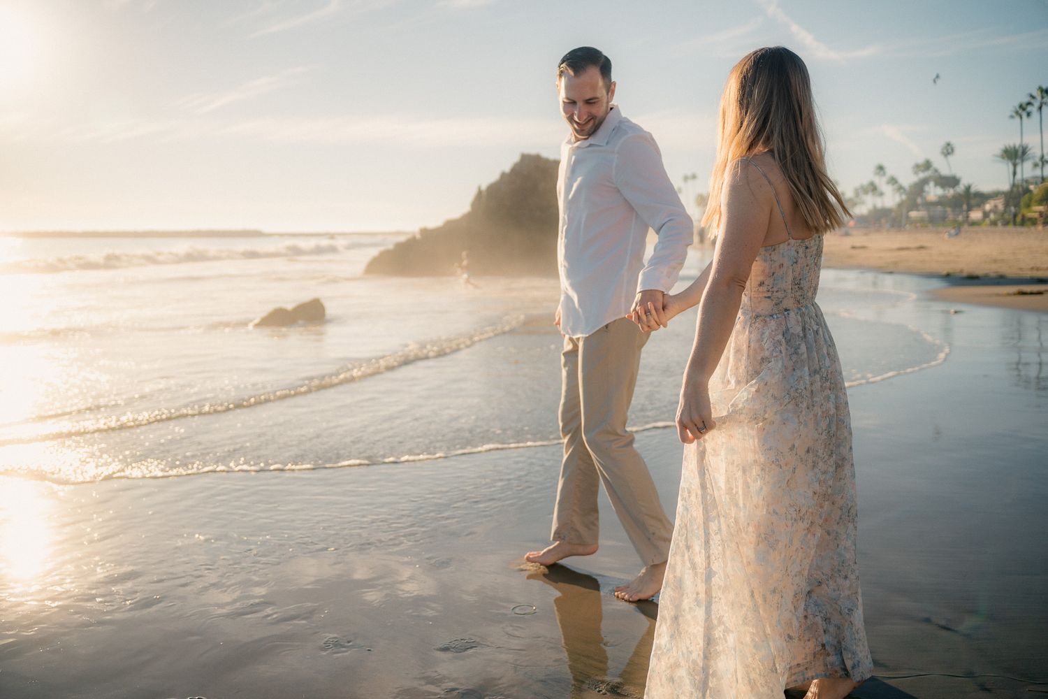 A couple holds hands while walking on a beach at sunset with ocean waves and rocks in the background.