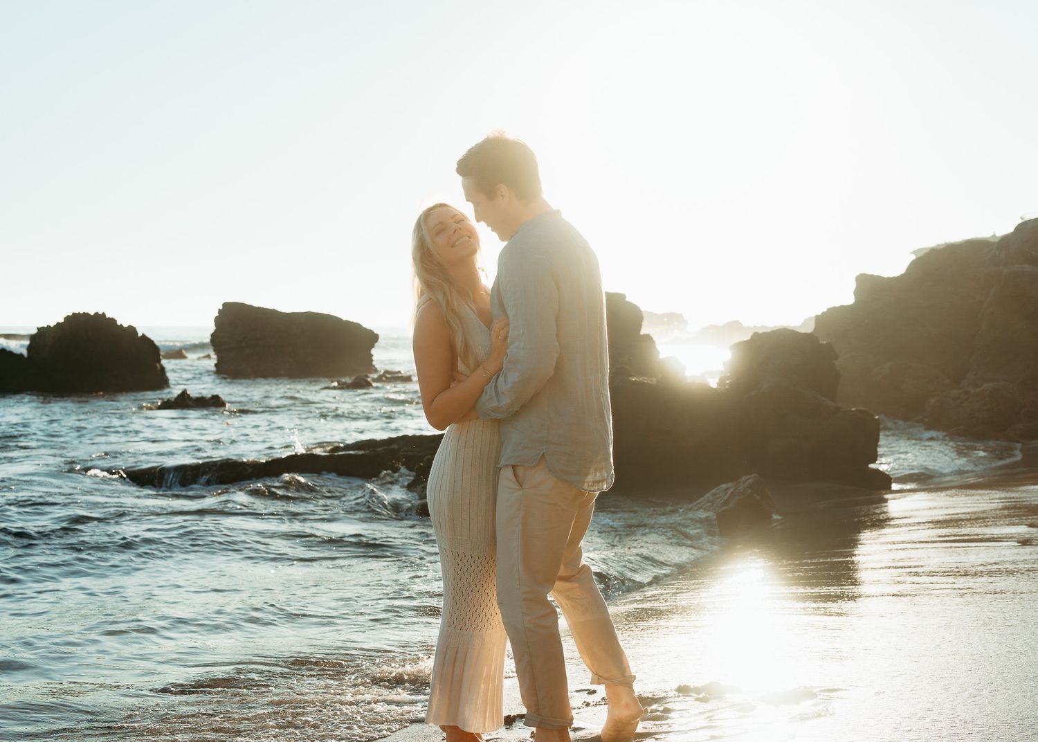 Intimate beachside moment captured during sunset with coastal rocks and gentle waves in the background.