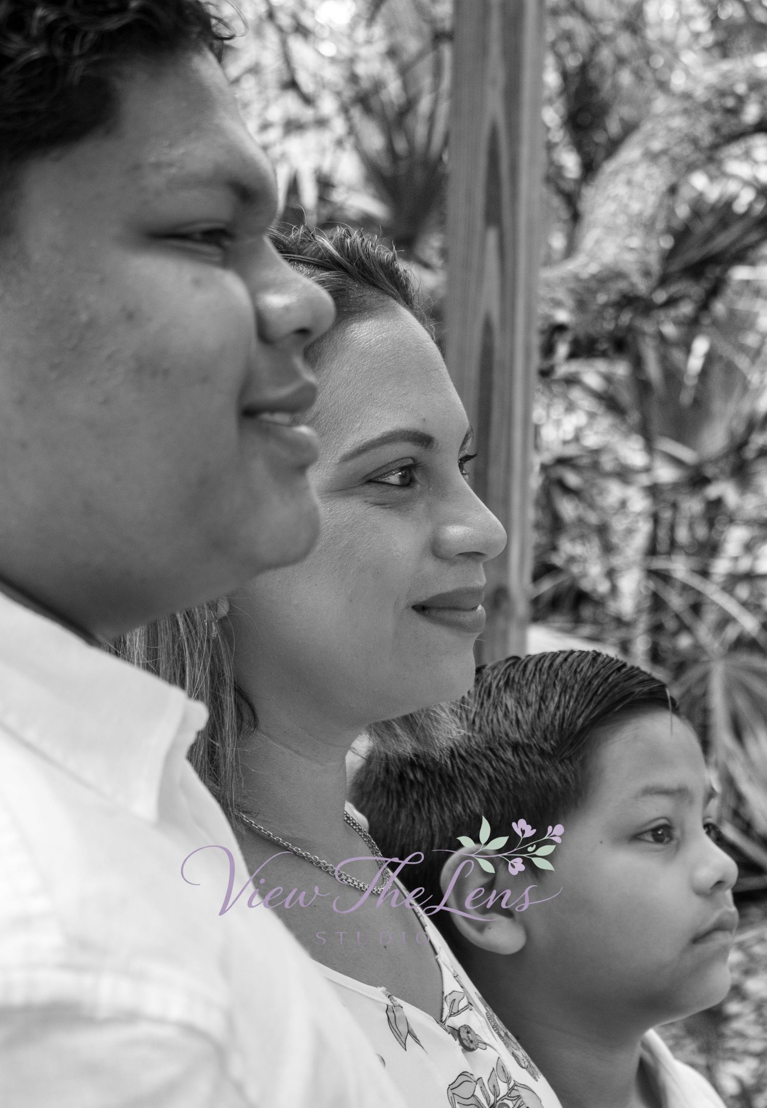 Black and white side-profile portrait of a parent and two children standing together outdoors with trees and foliage in the background.