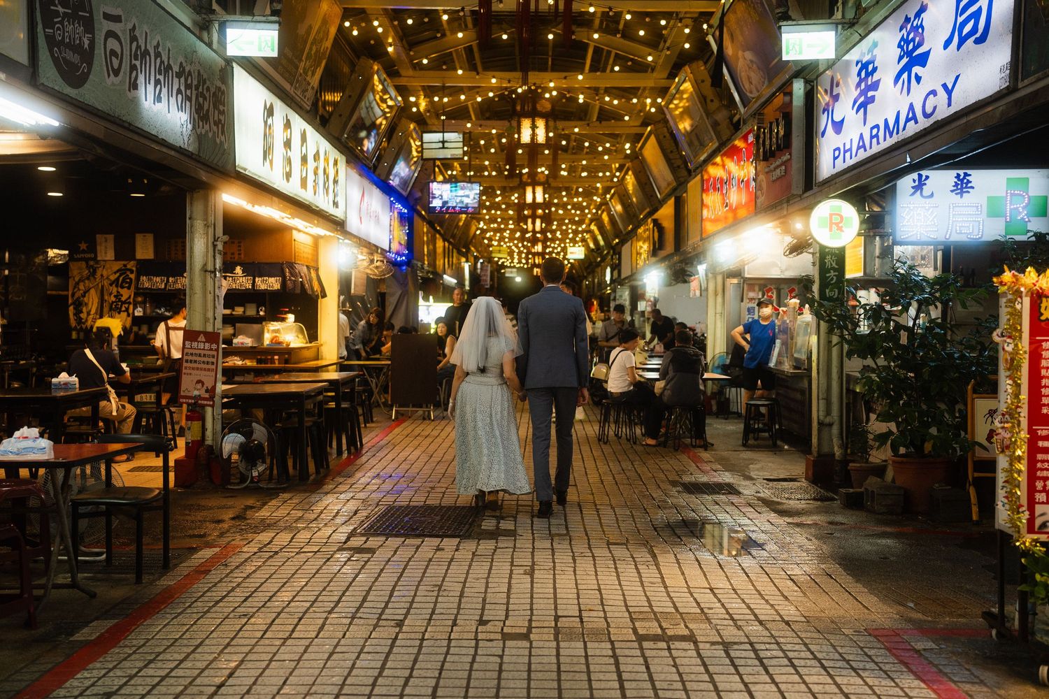 Bride and groom walking through Huaxi Street Night Market surrounded by local restaurants and glowing signs in Taipei.