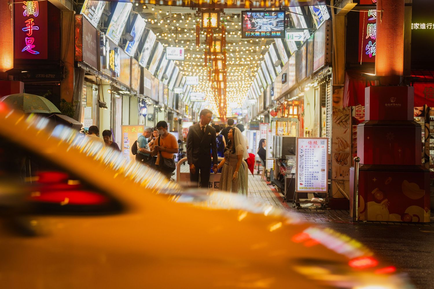 Taipei pre-wedding couple framed by a passing yellow taxi inside the colorful Huaxi Street Night Market.