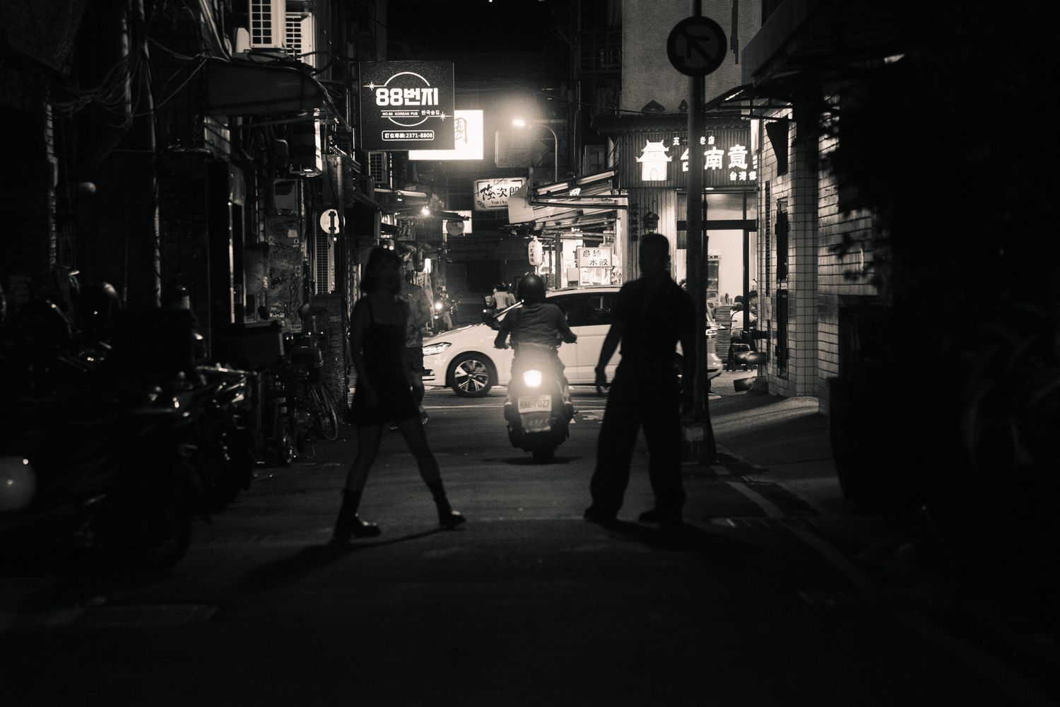 Black and white photo of a couple in a Ximending alley with a scooter passing between them at night