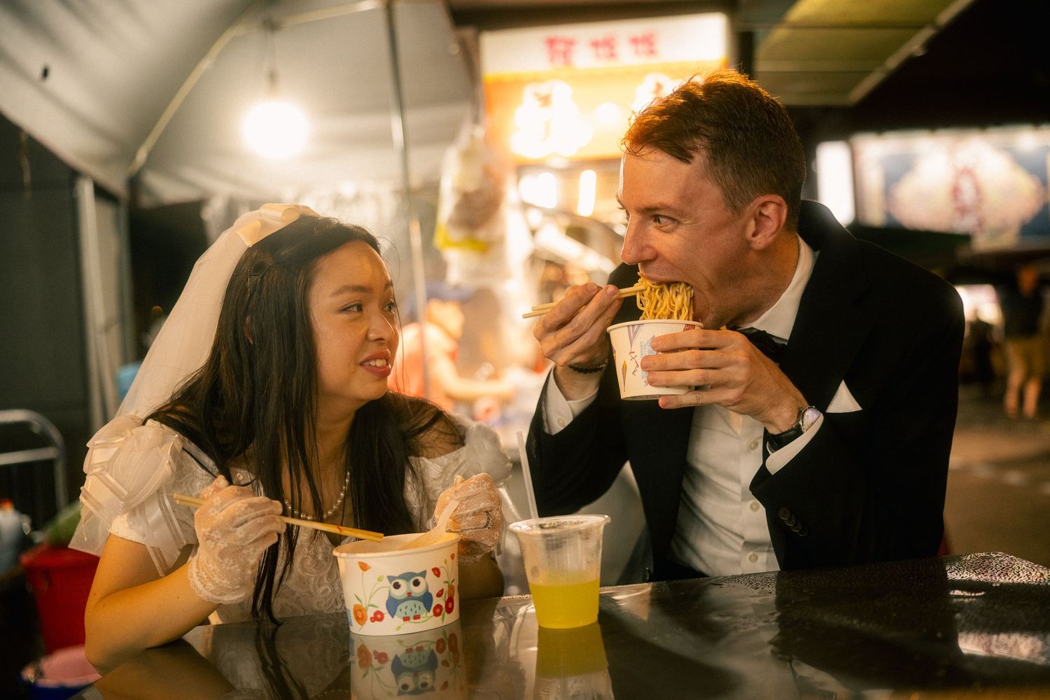 Bride watching her groom eat noodles at a Taiwanese street food stall during their Taipei pre-wedding photoshoot