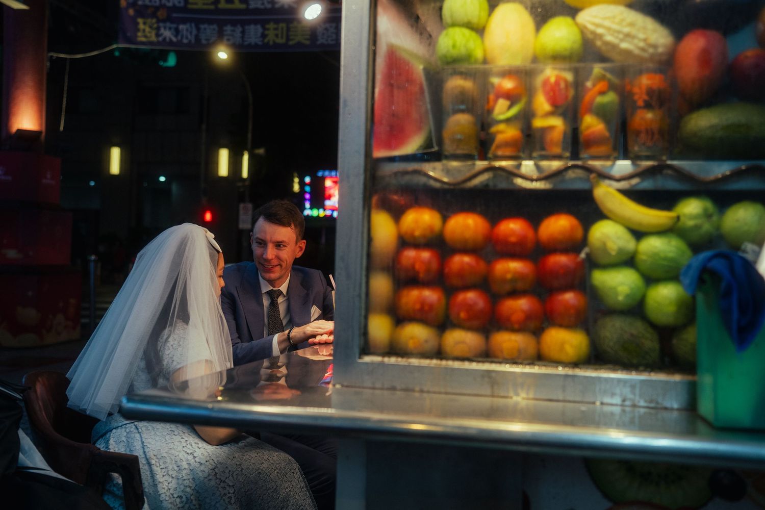 Candid couple portrait beside a traditional fruit stand during a nighttime pre-wedding session at Huaxi Street Night Market in Taipei.