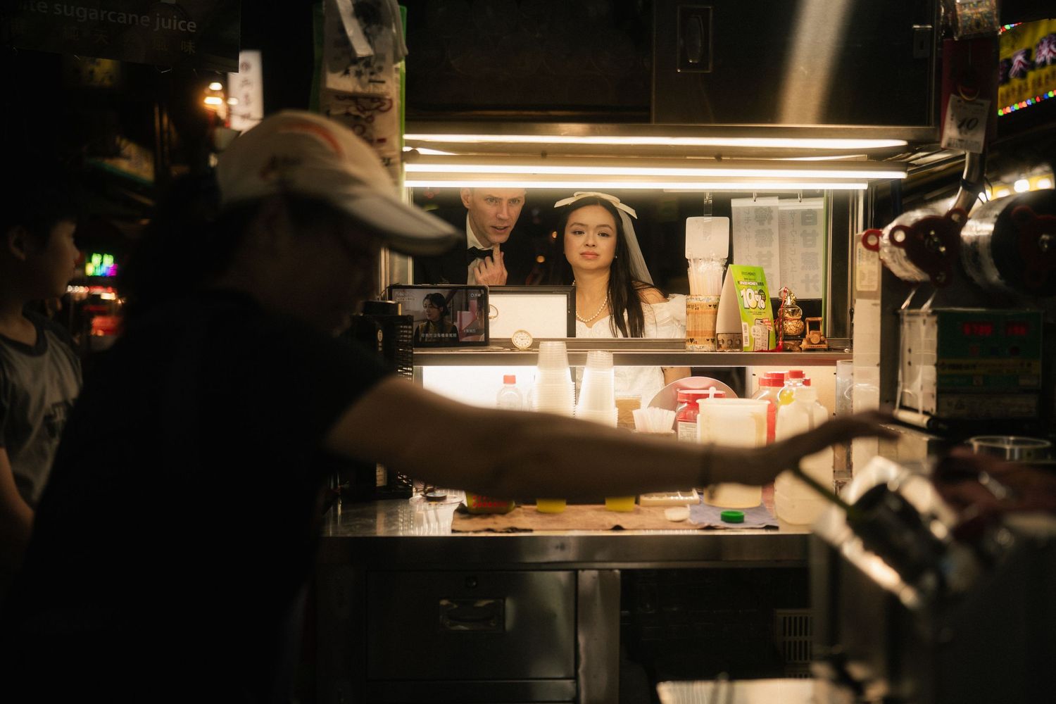 Bride and groom framed through a busy Taiwanese drink stall during a documentary-style Taipei photoshoot