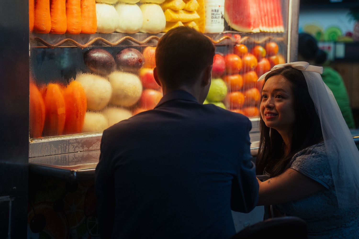 Bride smiling at her partner beside a colorful fruit stand during a cinematic Taipei night market pre-wedding photoshoot.