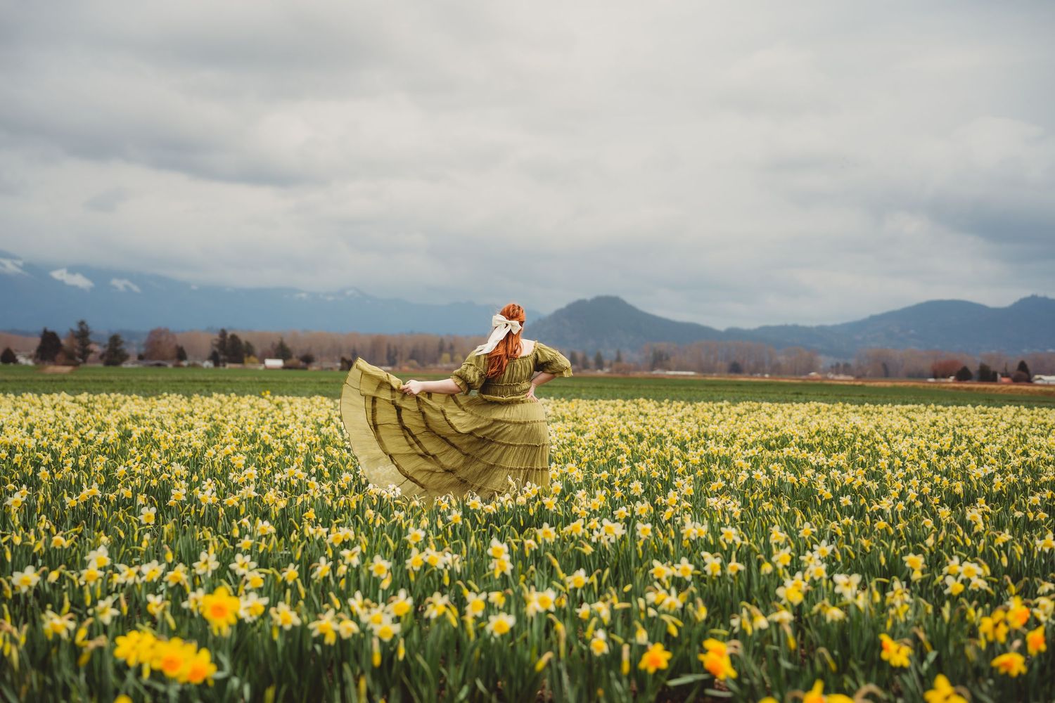 Woman in flowing dress twirling in a vibrant daffodil field with mountains and cloudy sky in background.