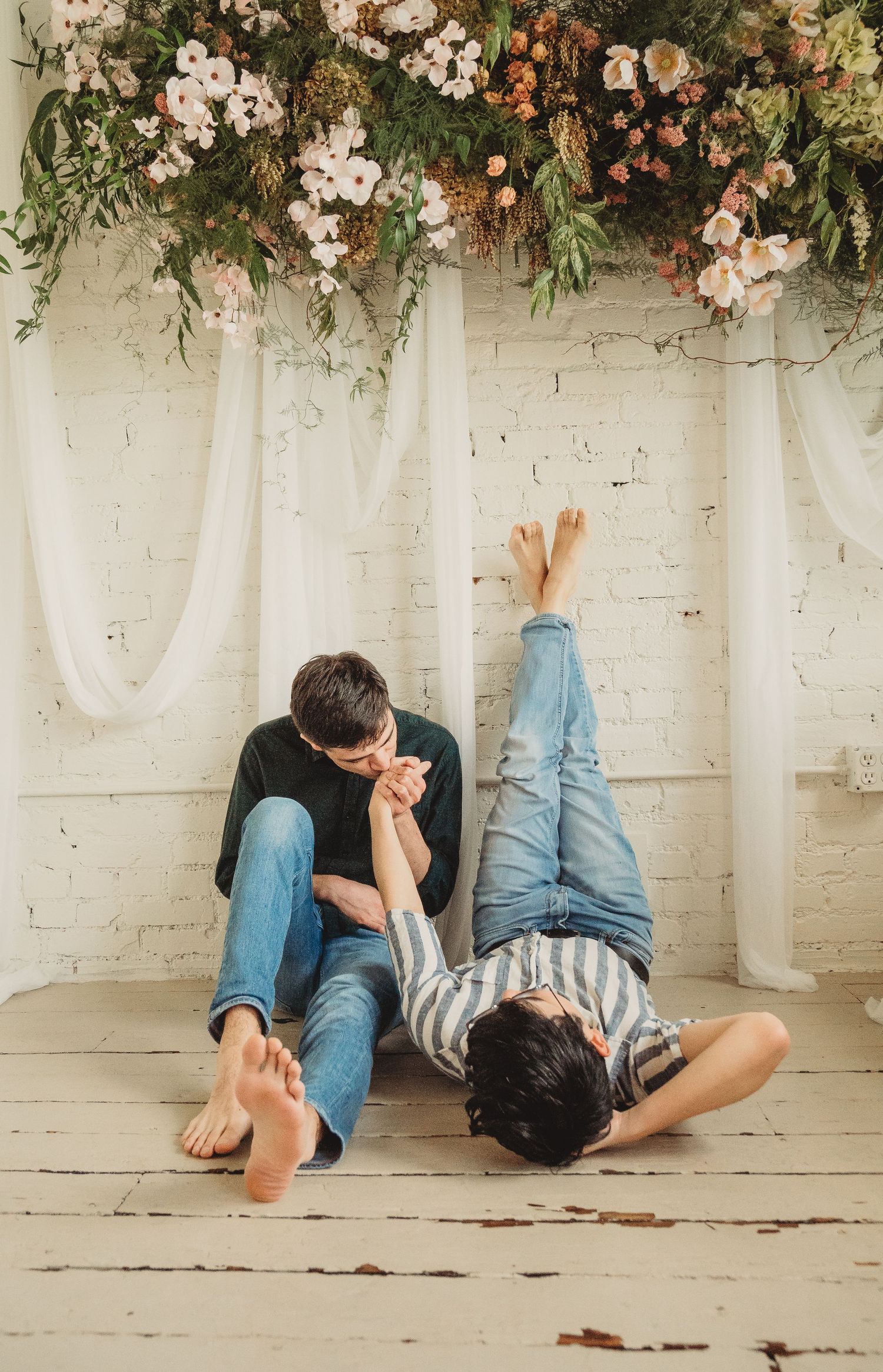 Two barefoot people sitting on a wooden floor holding hands beneath a floral arrangement with white draping backdrop.