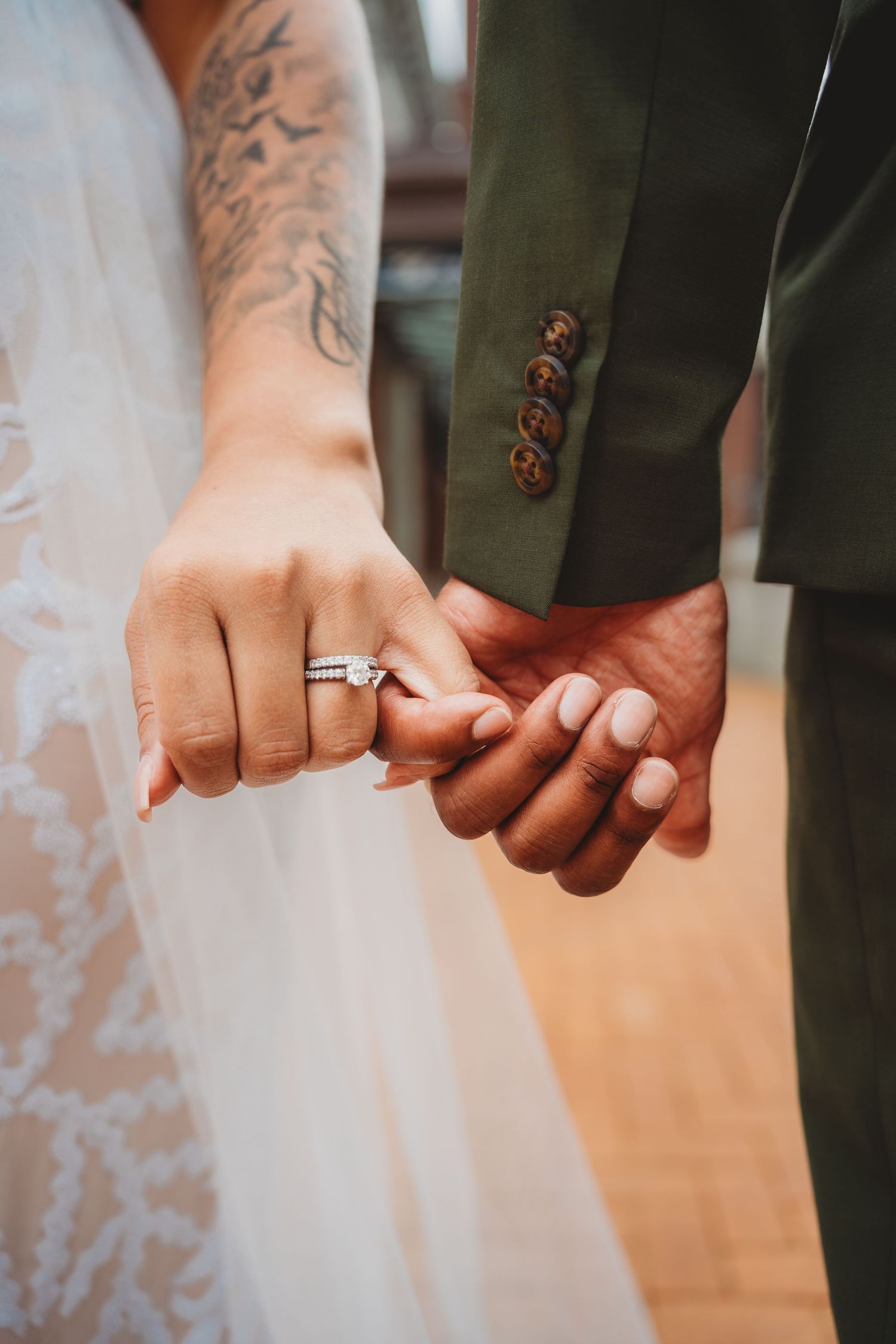 Bride and groom holding hands, showcasing a diamond engagement ring and wedding band, with lace dress and green suit.