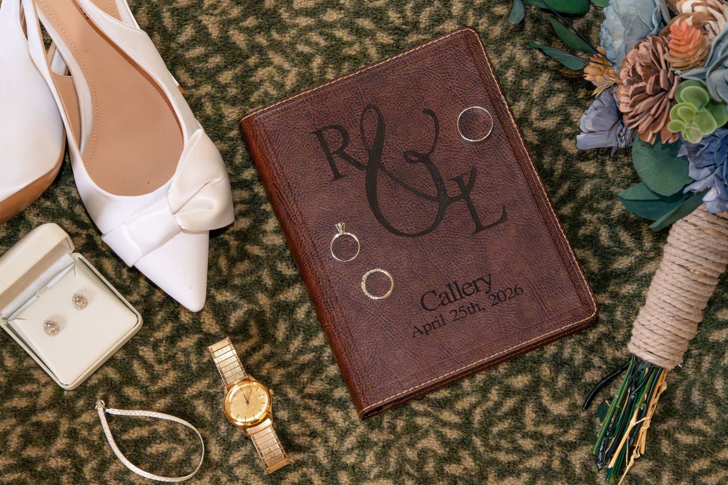 Elegant wedding flat lay with leather monogrammed album, gold rings, bridal heels, watch, earrings, and floral bouquet.