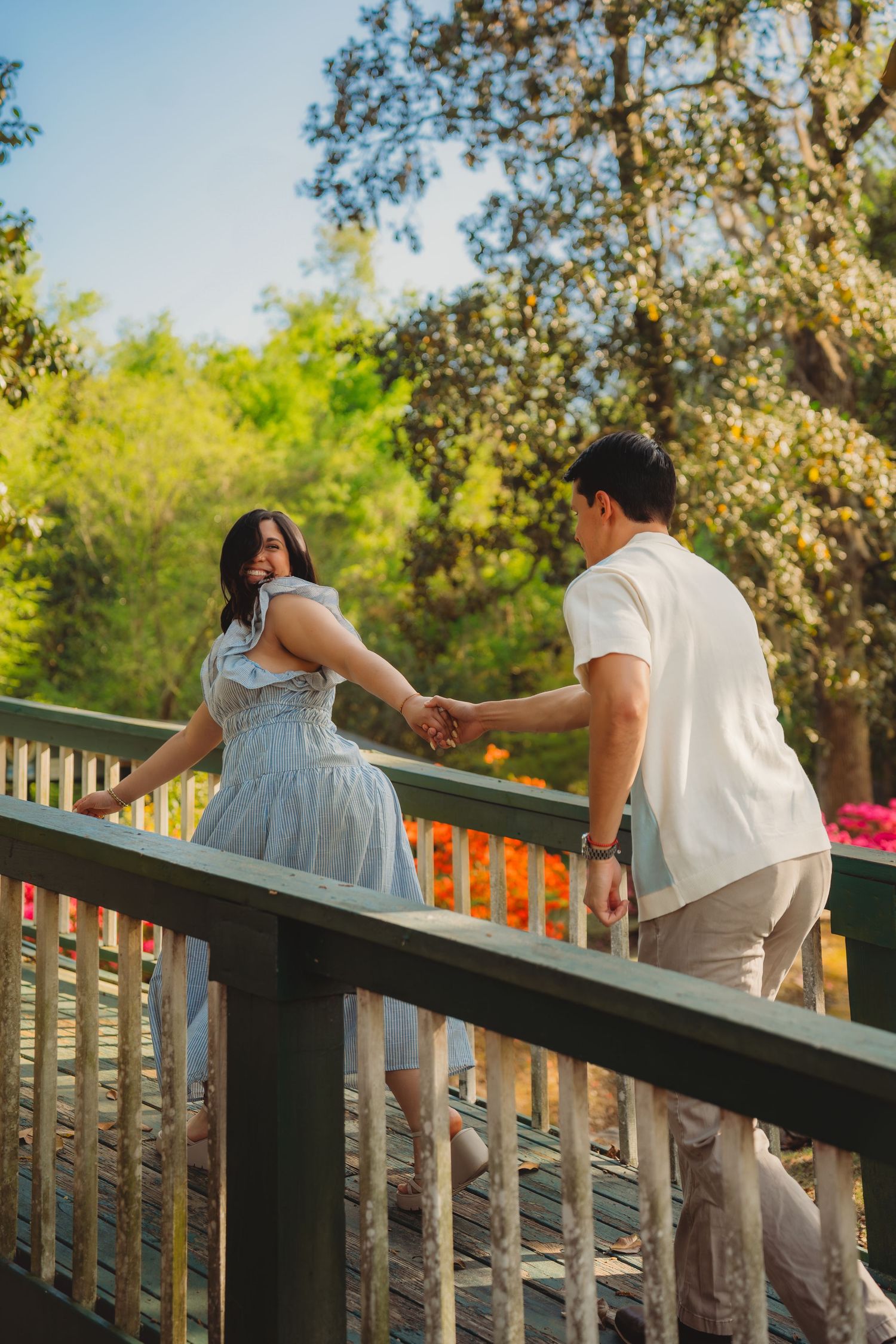 Couple holding hands on a wooden bridge surrounded by lush green trees on a sunny day.