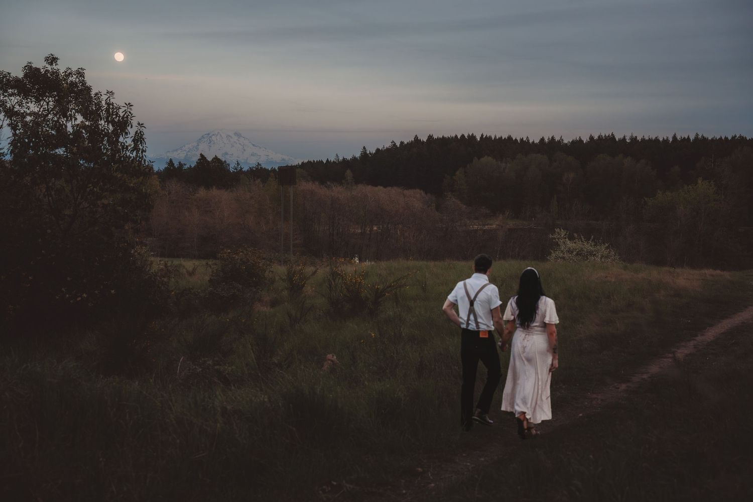 Couple walking hand in hand through a moonlit forest meadow at dusk, dressed in wedding attire.