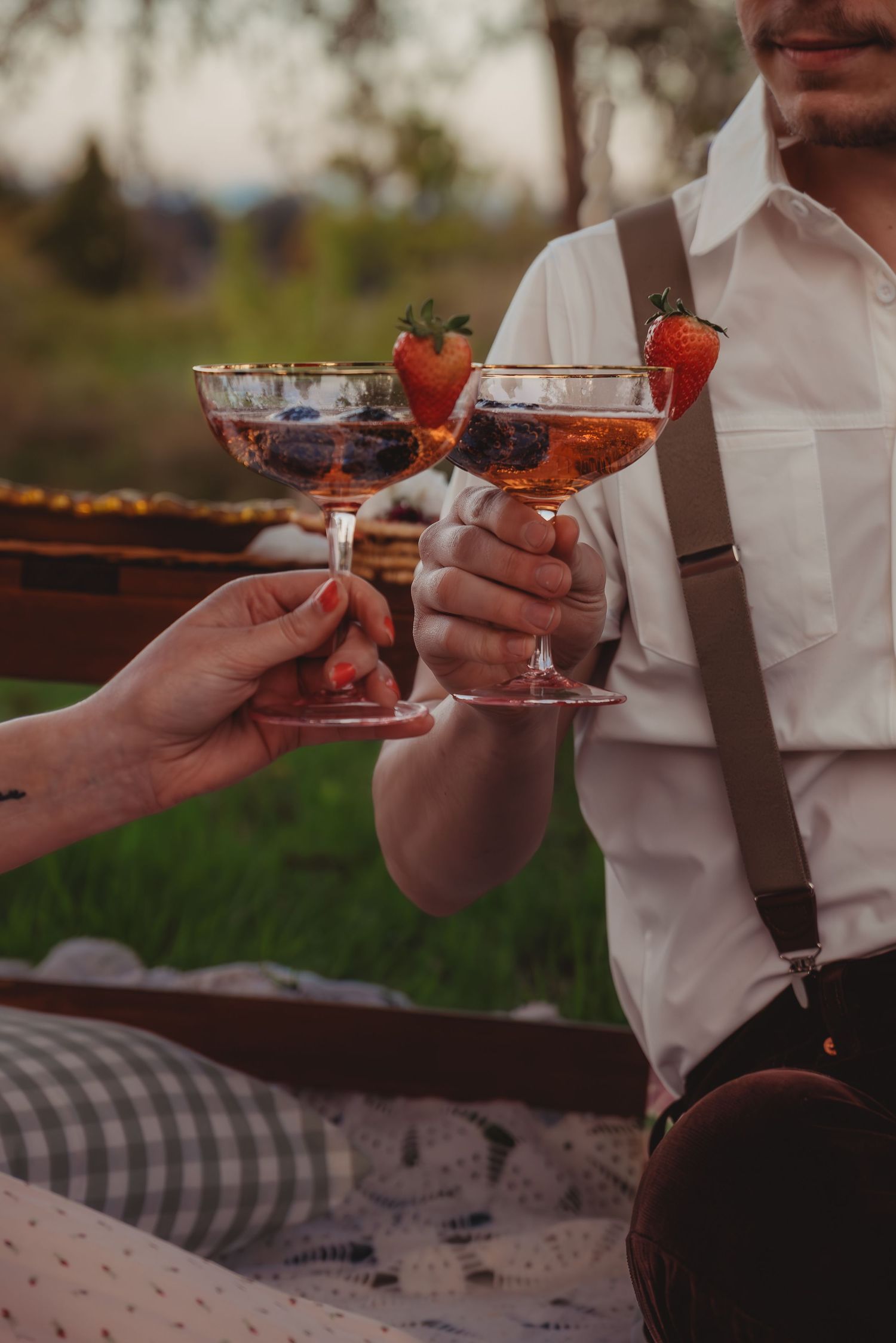 Couple toasting with elegant champagne coupe glasses garnished with fresh strawberries at an outdoor picnic.