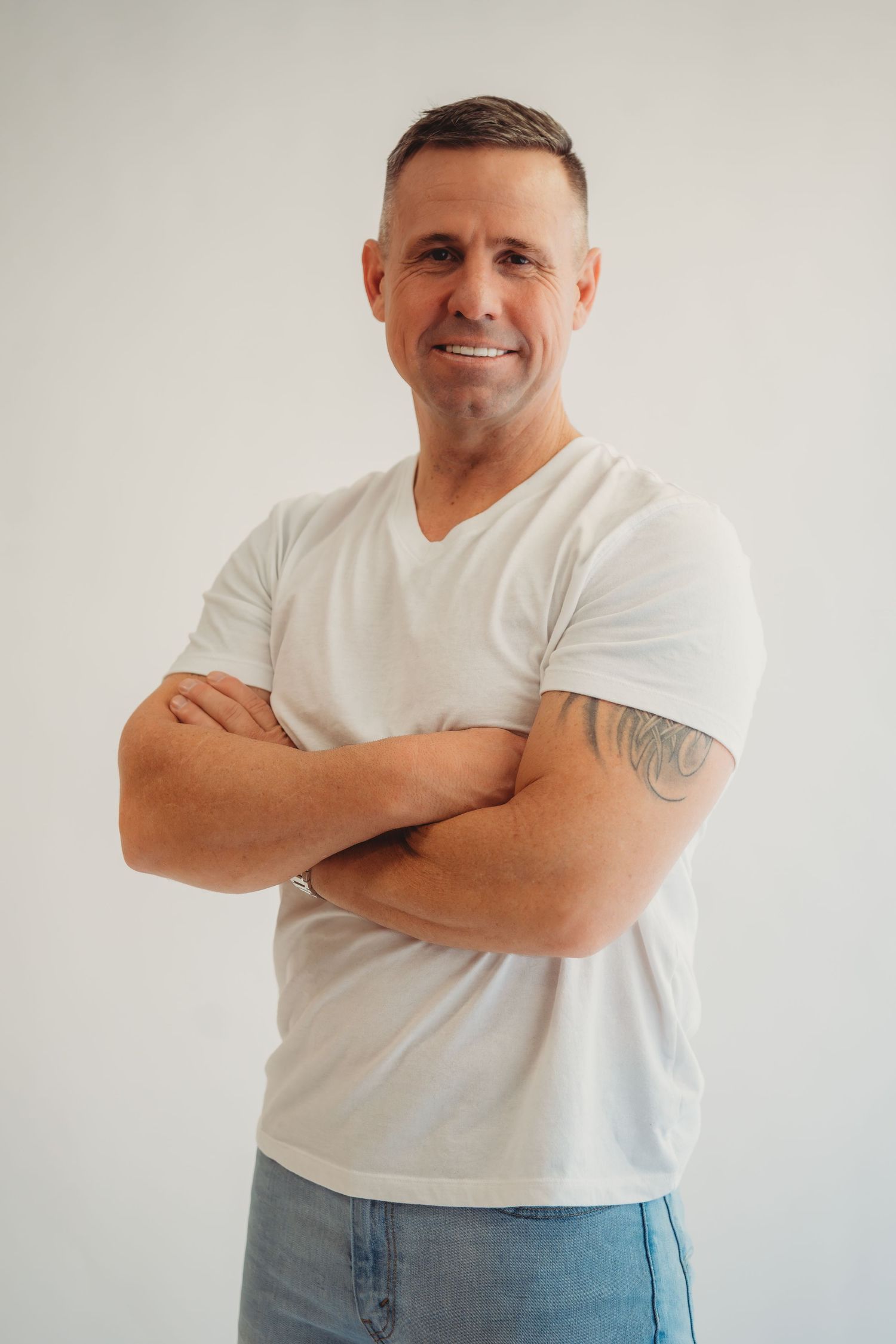 Smiling middle-aged man with tattoo on arm, wearing white v-neck t-shirt and jeans, standing with arms crossed.