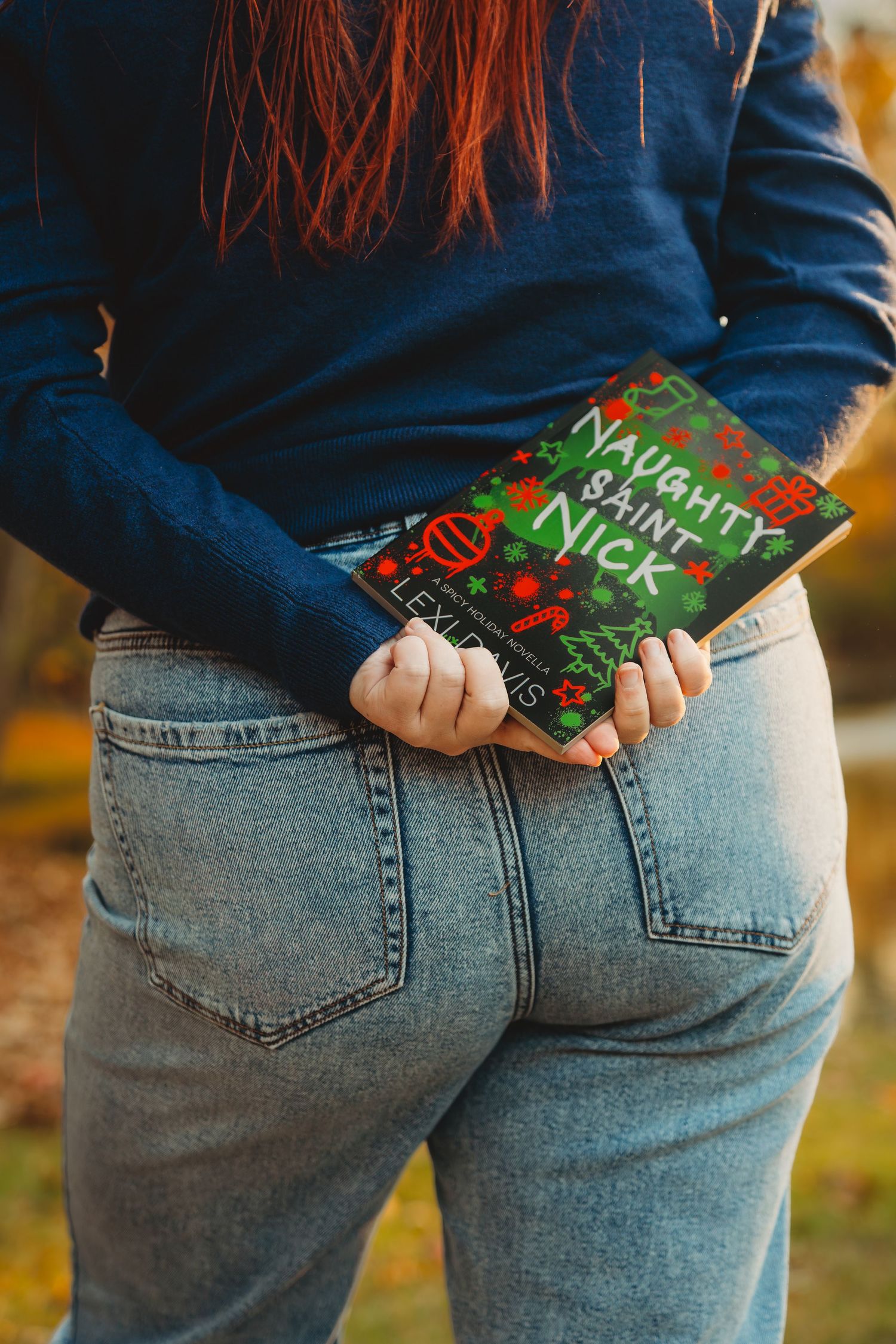 Woman in jeans holding a Naughty Saint Nick holiday book behind her back outdoors.