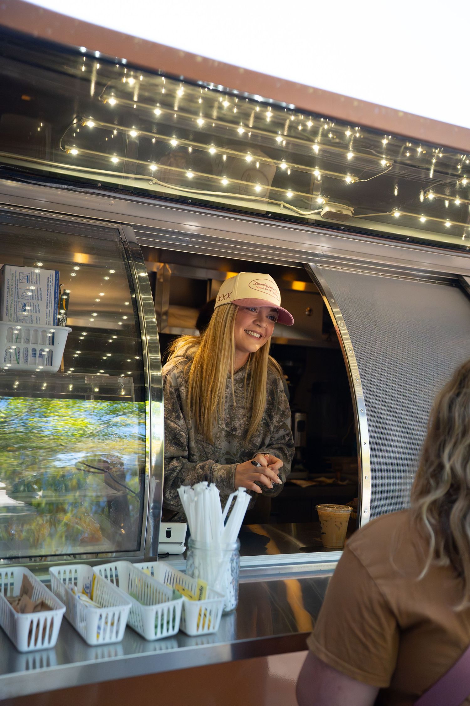 Smiling blonde woman in camo jacket serving customers from a decorated food truck window. West Ashley South Carolina