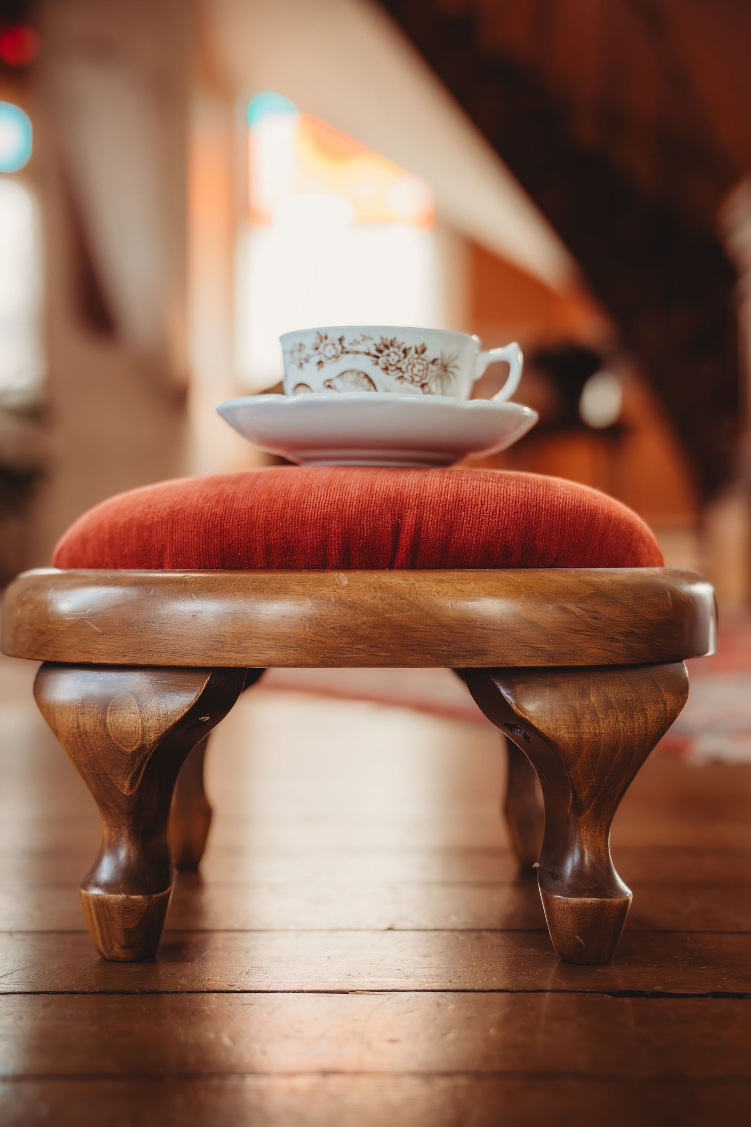 Antique wooden footstool with red velvet cushion holding a delicate floral teacup and saucer on hardwood floor.