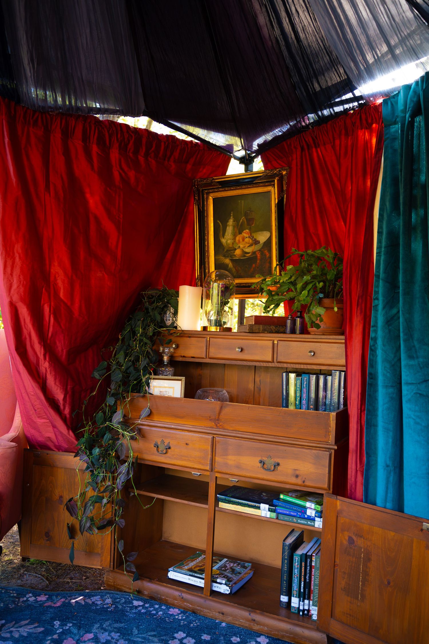 Wooden cabinet with books, candles and trailing plants beneath a framed painting, set in a cozy tent with red and teal velvet curtains.