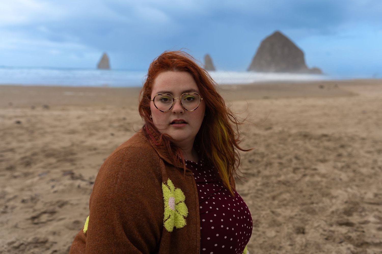 Woman with red hair and glasses stands on a sandy beach with Haystack Rock in the background under cloudy skies.