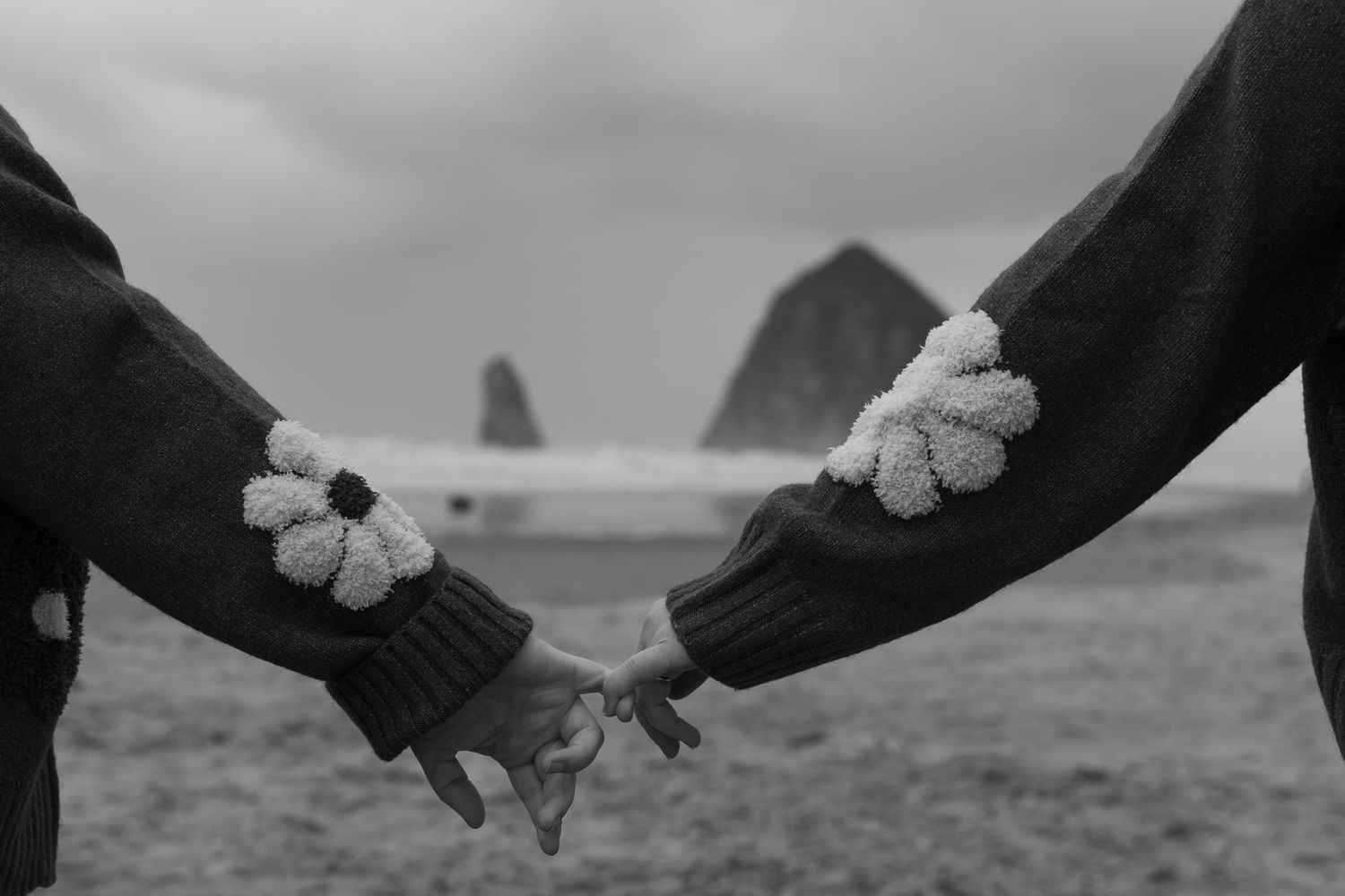 Couple holding hands in matching knit sweaters with floral cuffs at Cannon Beach with Haystack Rock backdrop.