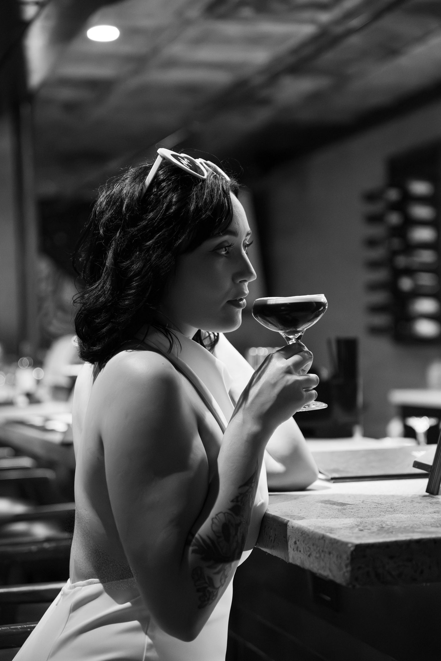 Black and white photo of a tattooed woman with sunglasses on her head sipping a cocktail at a stylish bar counter.
