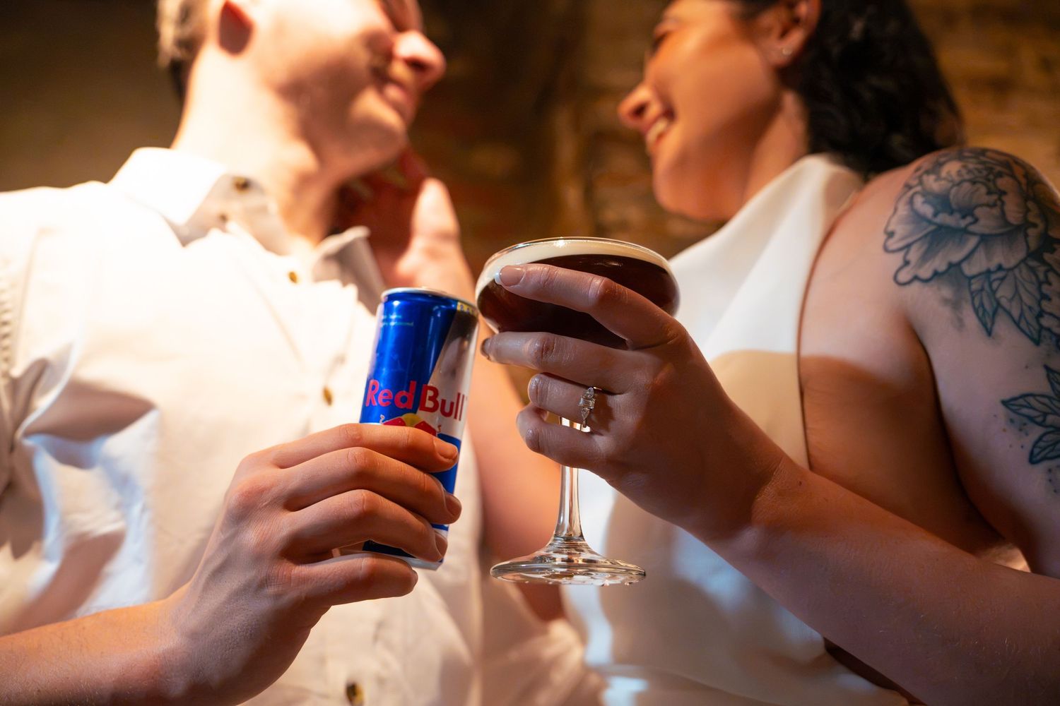 Two smiling people toast with a blue and silver energy drink can and a glass of red wine in a warm, cozy venue.