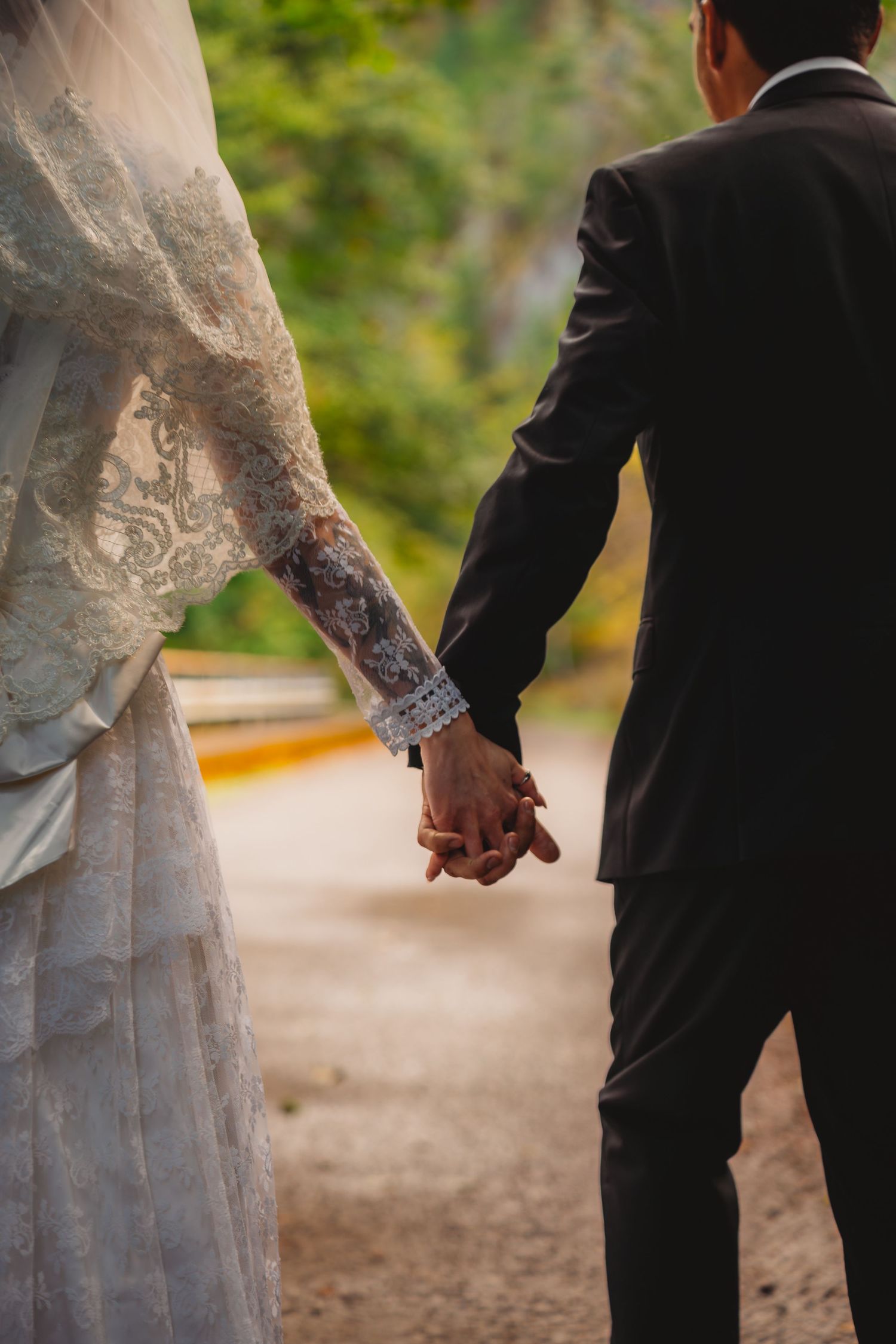 Bride and groom holding hands from behind, bride in lace wedding dress, groom in black suit, walking outdoors.