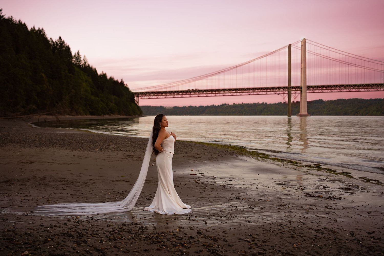 Bride in white gown with long veil stands on rocky shore at sunset with suspension bridge in background.