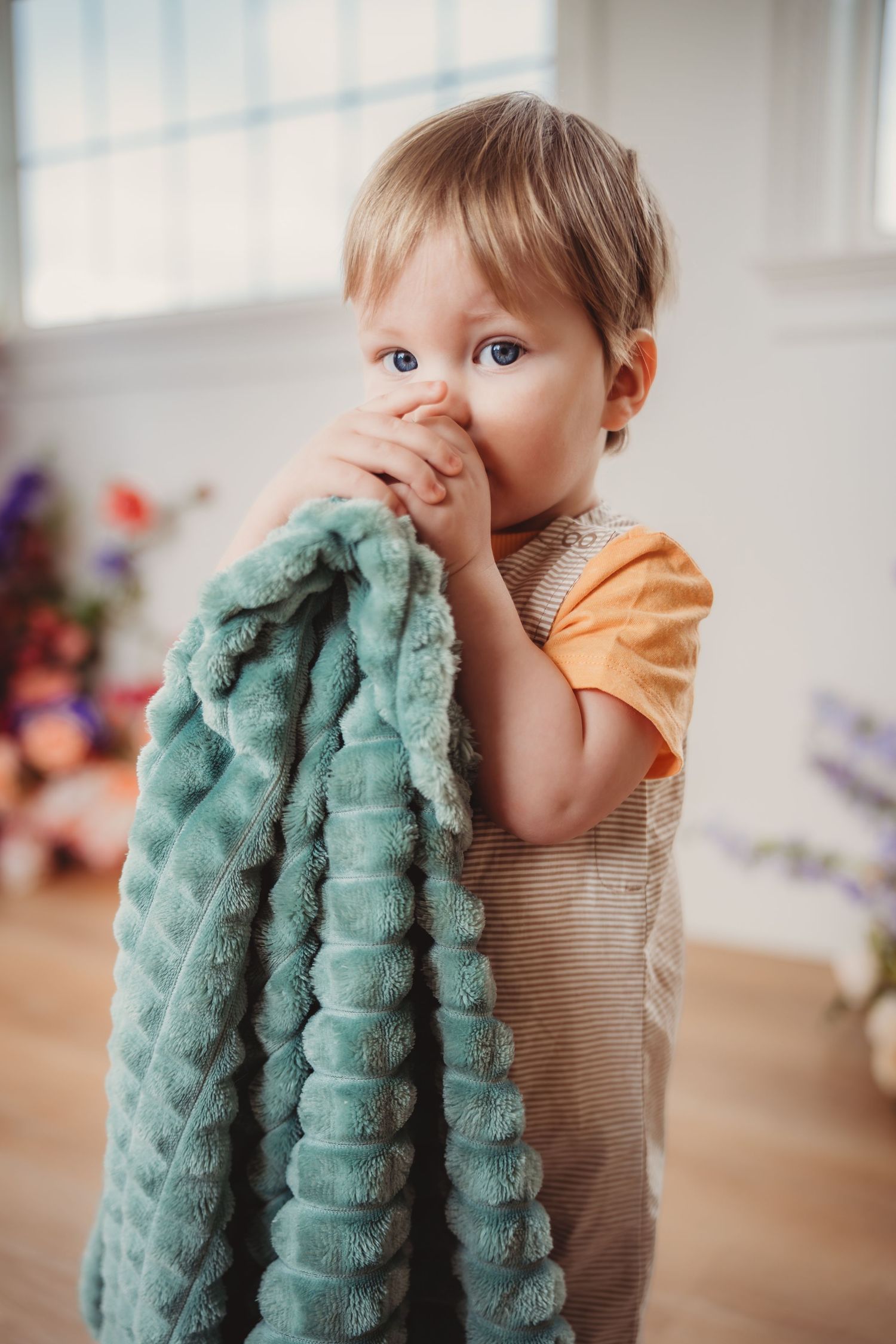 Adorable toddler in orange shirt holding teal blanket indoors near window. studio South Carolina