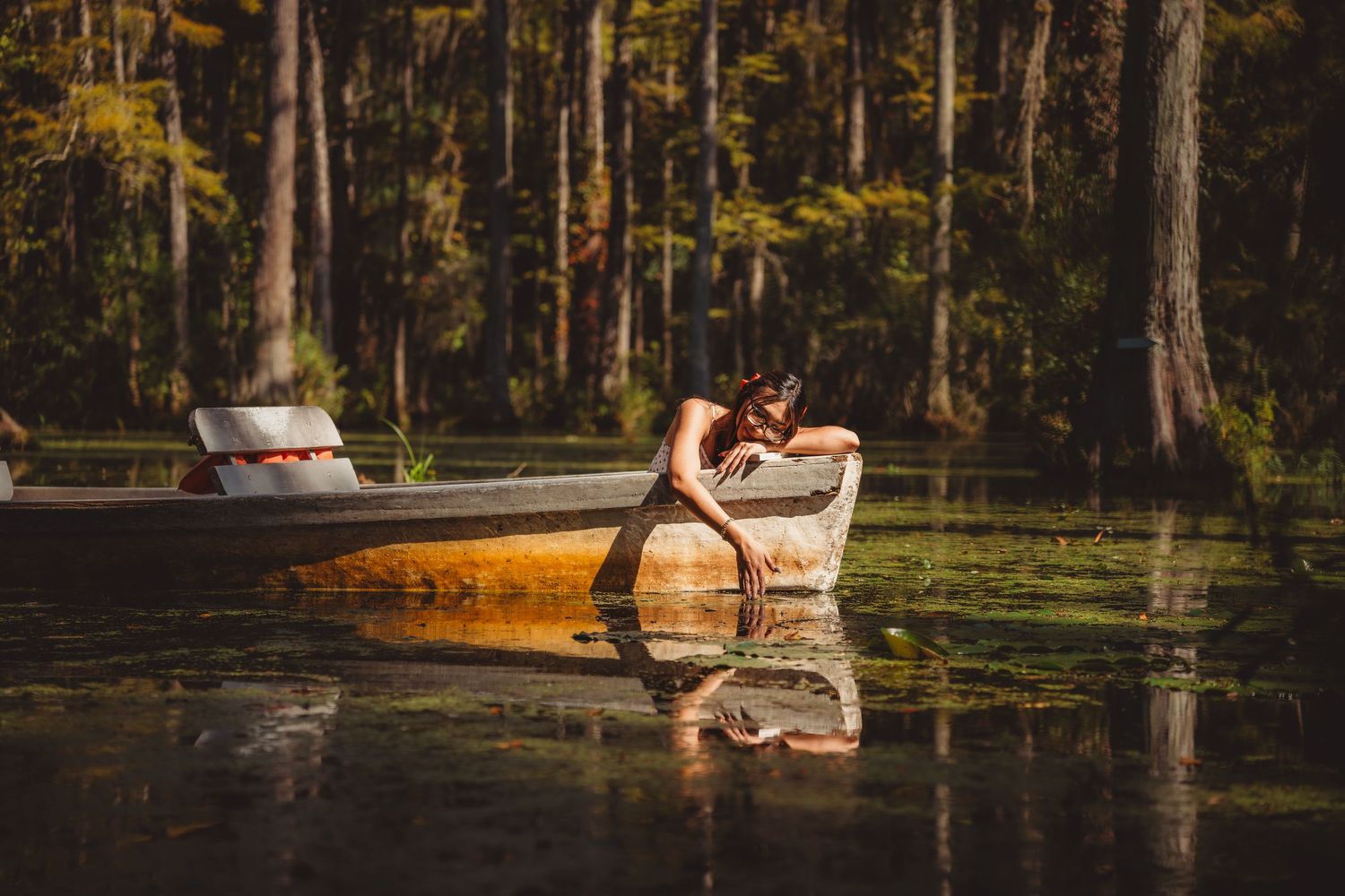 Woman leaning over edge of wooden boat on a serene cypress swamp, golden light reflecting on still water.