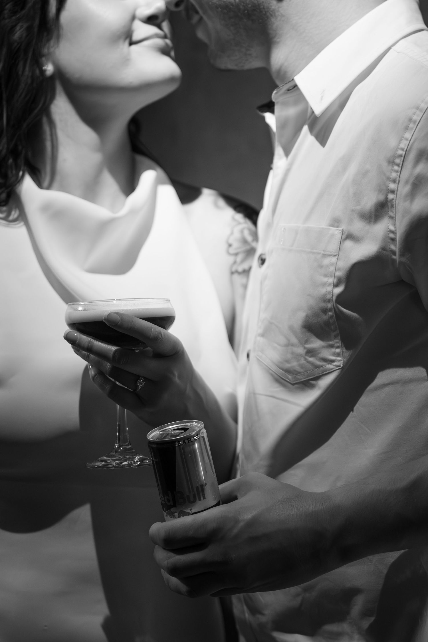 Black and white photo of a smiling woman holding a cocktail glass leaning in to kiss a man holding a beverage can.