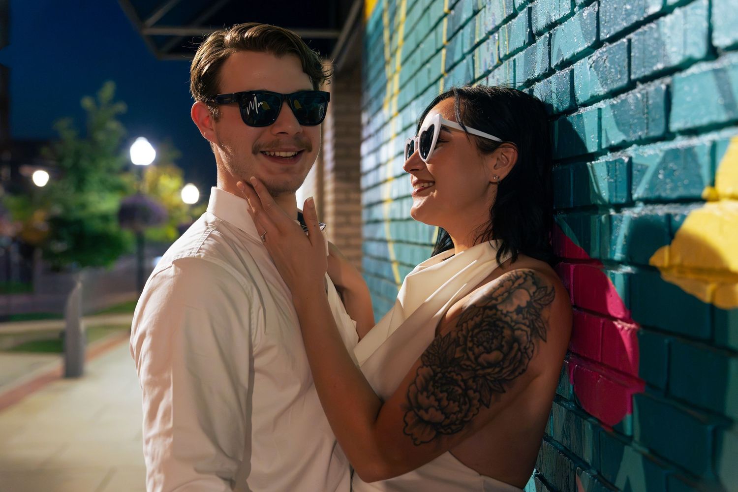Smiling couple in sunglasses embrace beside a colorful graffiti brick wall on a city street at dusk, her arm showing a floral tattoo.