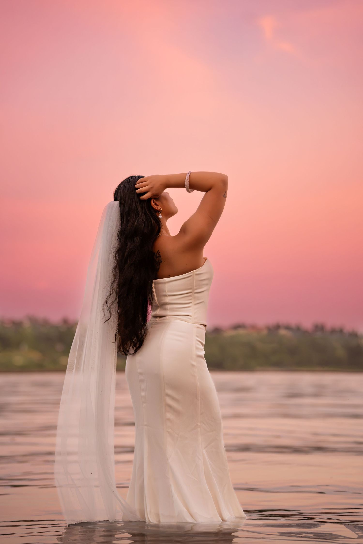 Bride in white gown and veil standing in water at pink sunset, viewed from behind.