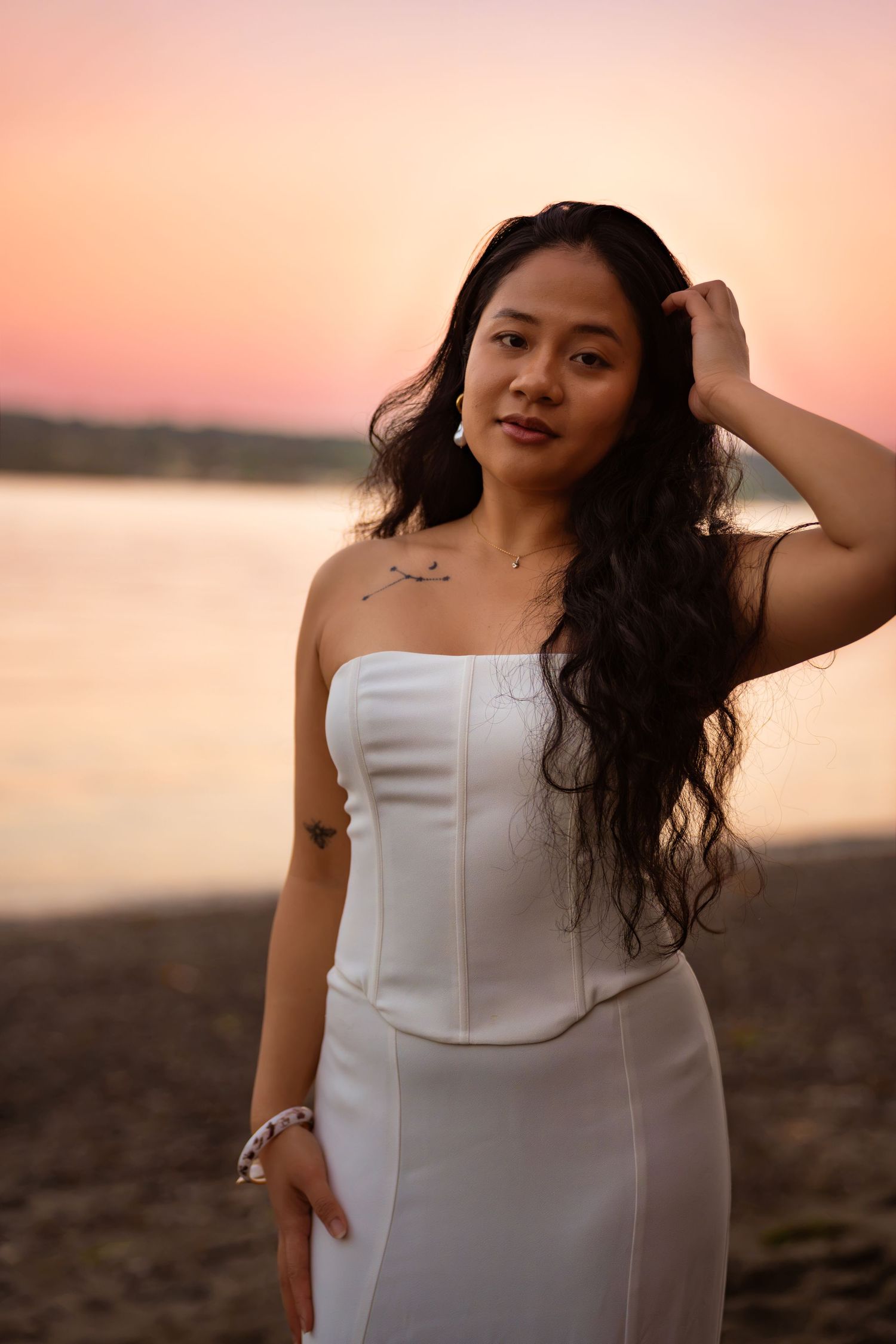 Woman in white strapless dress posing on beach at sunset with pink sky and water in background.